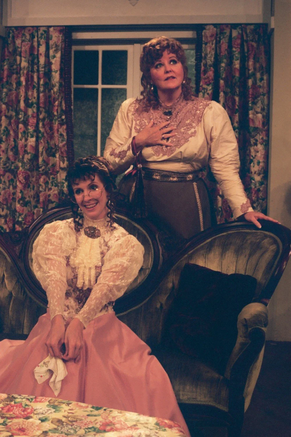 Two women in Victorian-style dresses, one sitting on a vintage chair and the other standing, indoors with floral curtains.