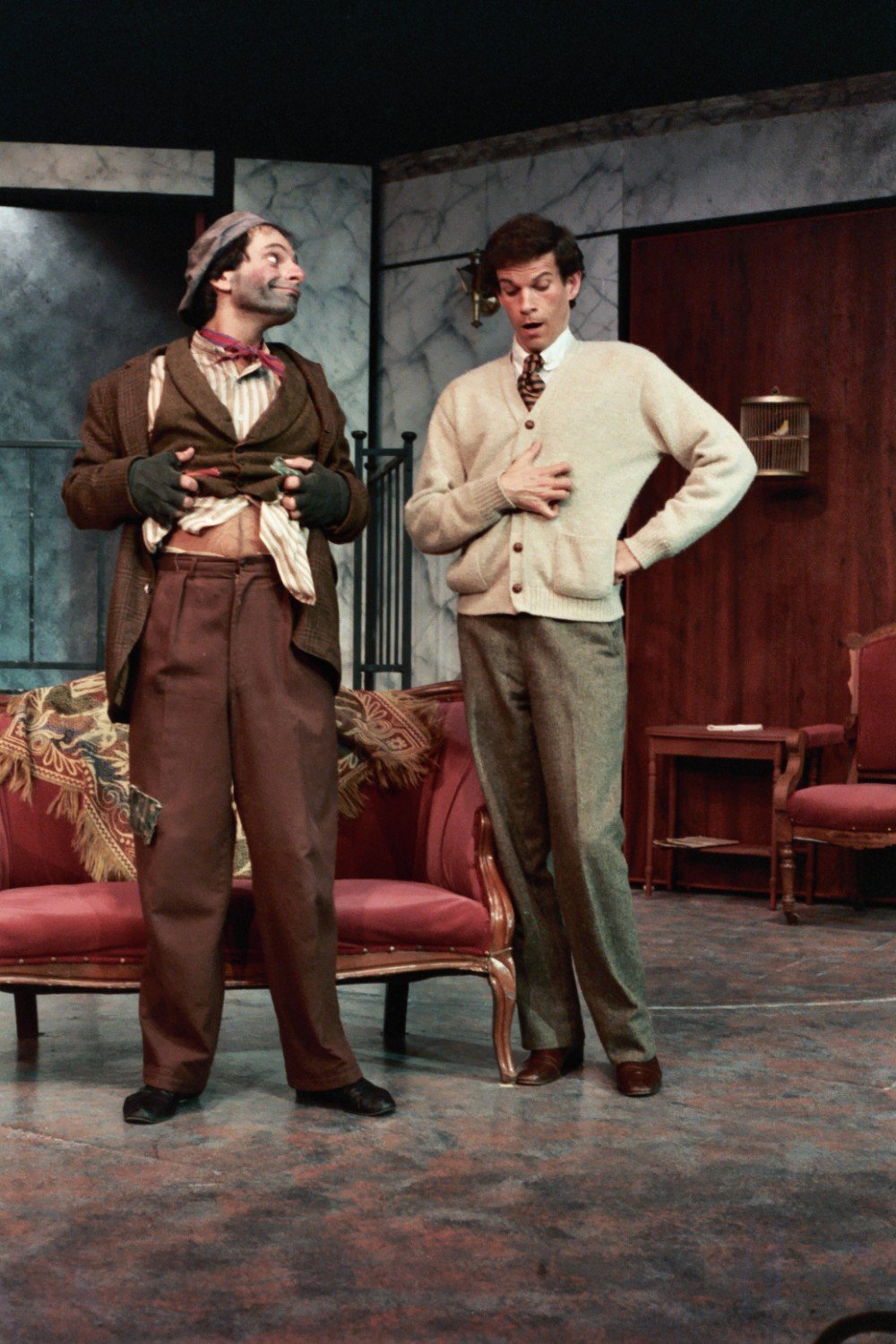 Two actors on stage, one dressed as a shabby, comical character, the other in a neat sweater and tie, in a theatrical setting with vintage furniture and decor.