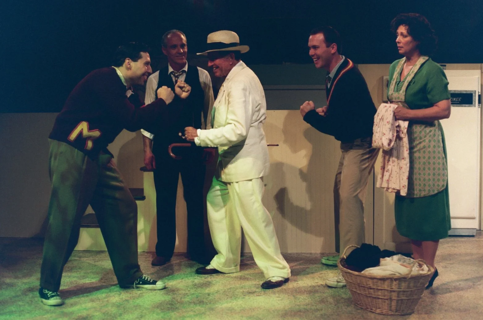 A group of five people on stage, engaged in a lively scene. Two men appear to be playfully sparring, one in a maroon sweater and sneakers, the other in a white suit and hat. A man in the background watches, wearing a vest and holding a cane. Another 