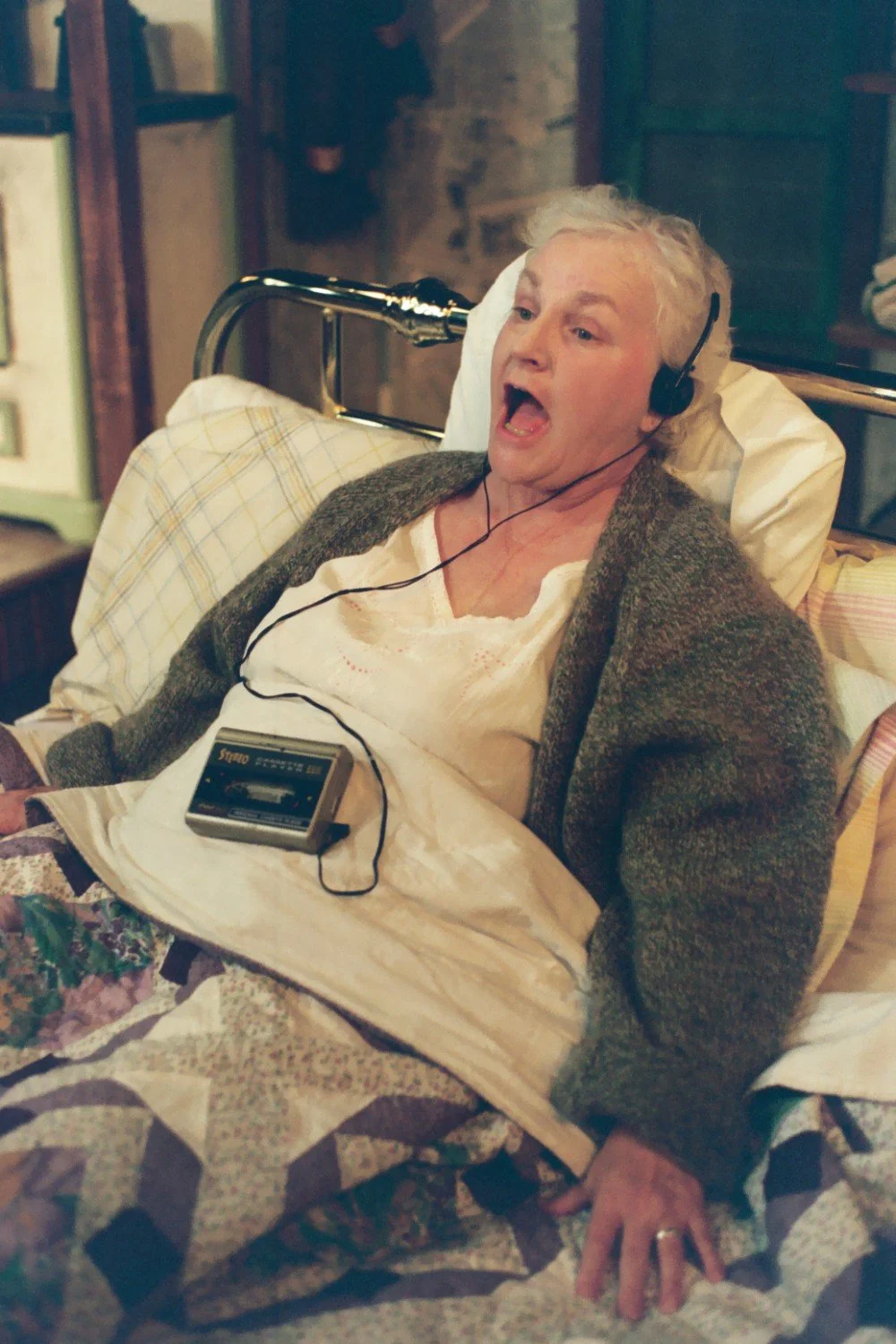 Older woman in bed listening to headphones, holding a cassette player, wearing a cardigan and nightgown.