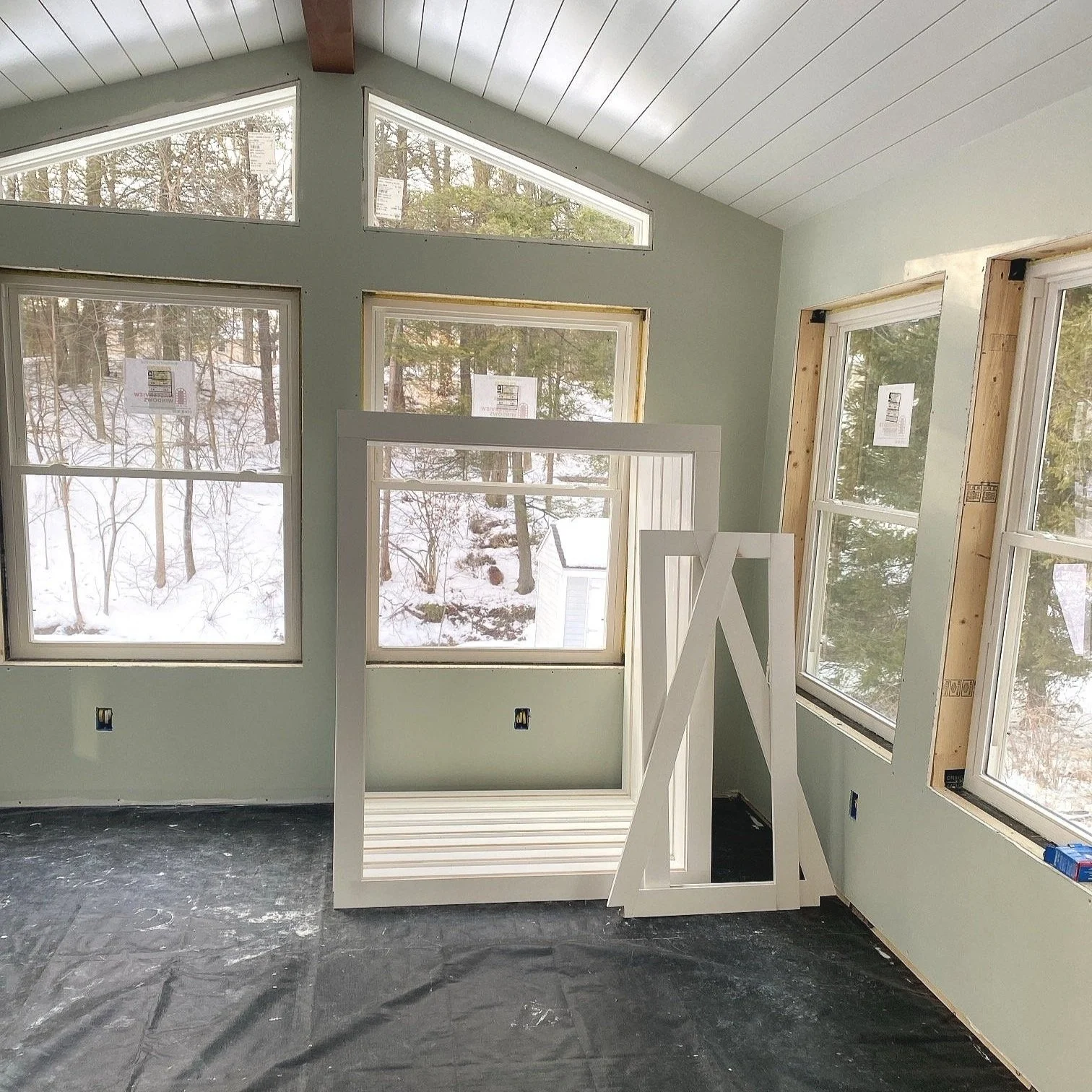 Interior of a room under construction with several windows and a vaulted ceiling, with construction materials and wooden windows trim frame leaning against the wall.