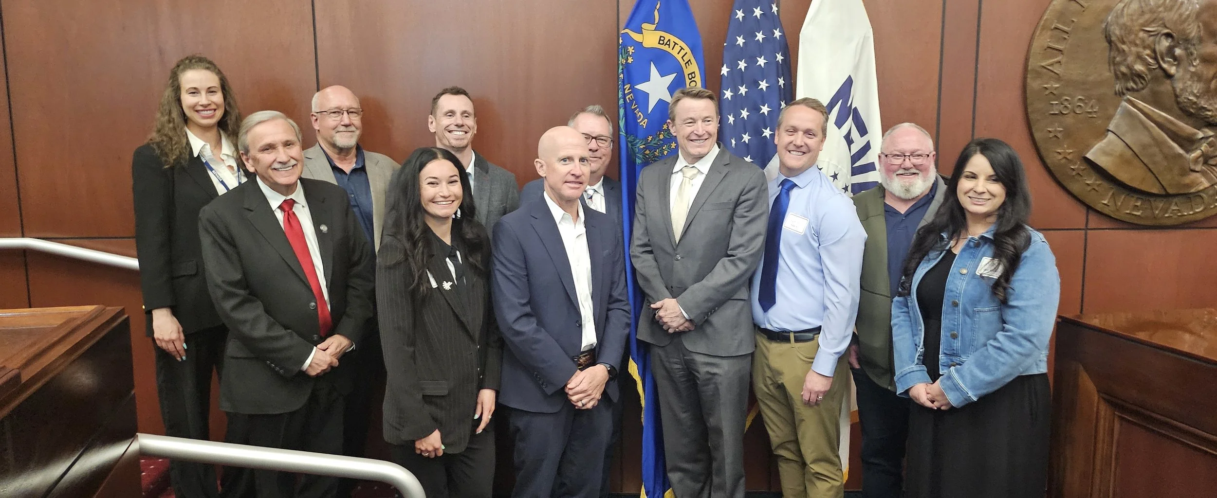 Group of ten people, including men and women, standing inside a room with historical wood-paneled walls and flags, smiling for a photo.