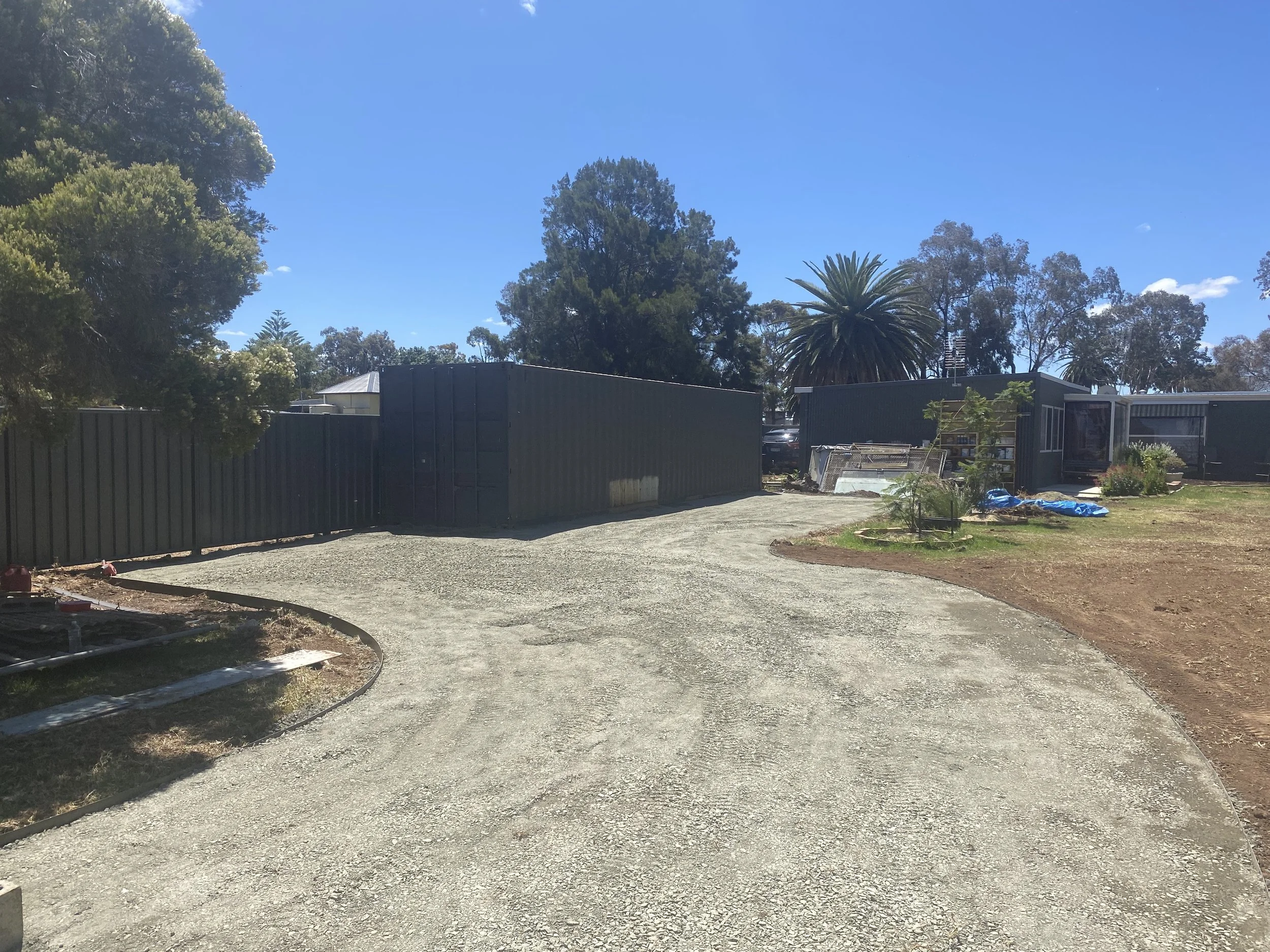 A gravel driveway curves to the right beside a fenced yard with trees and a small garden in a residential area on a sunny day.