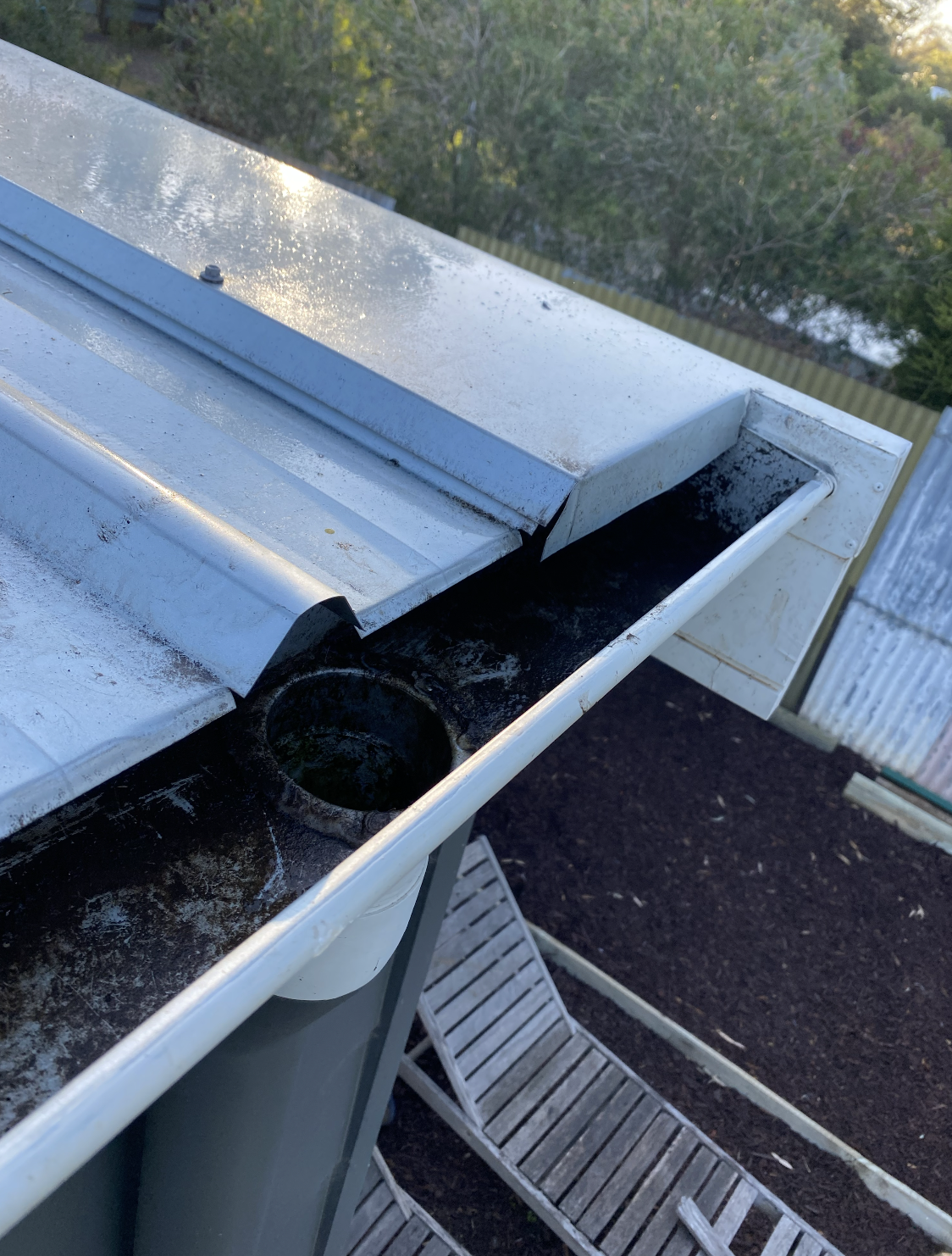 Close-up of a metal gutter on a house, showing accumulation of black debris and water in a circular section, with a background of trees and a dark ground.