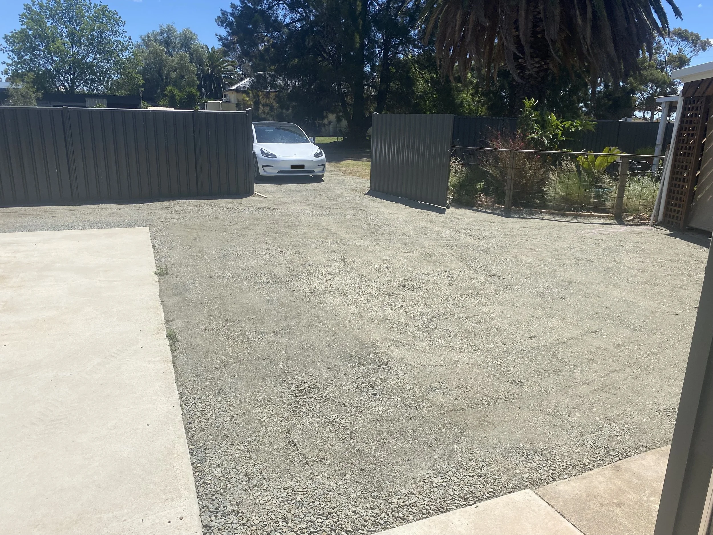 A driveway with a gravel surface, fenced on both sides, with a white car parked near the fence on the left side.