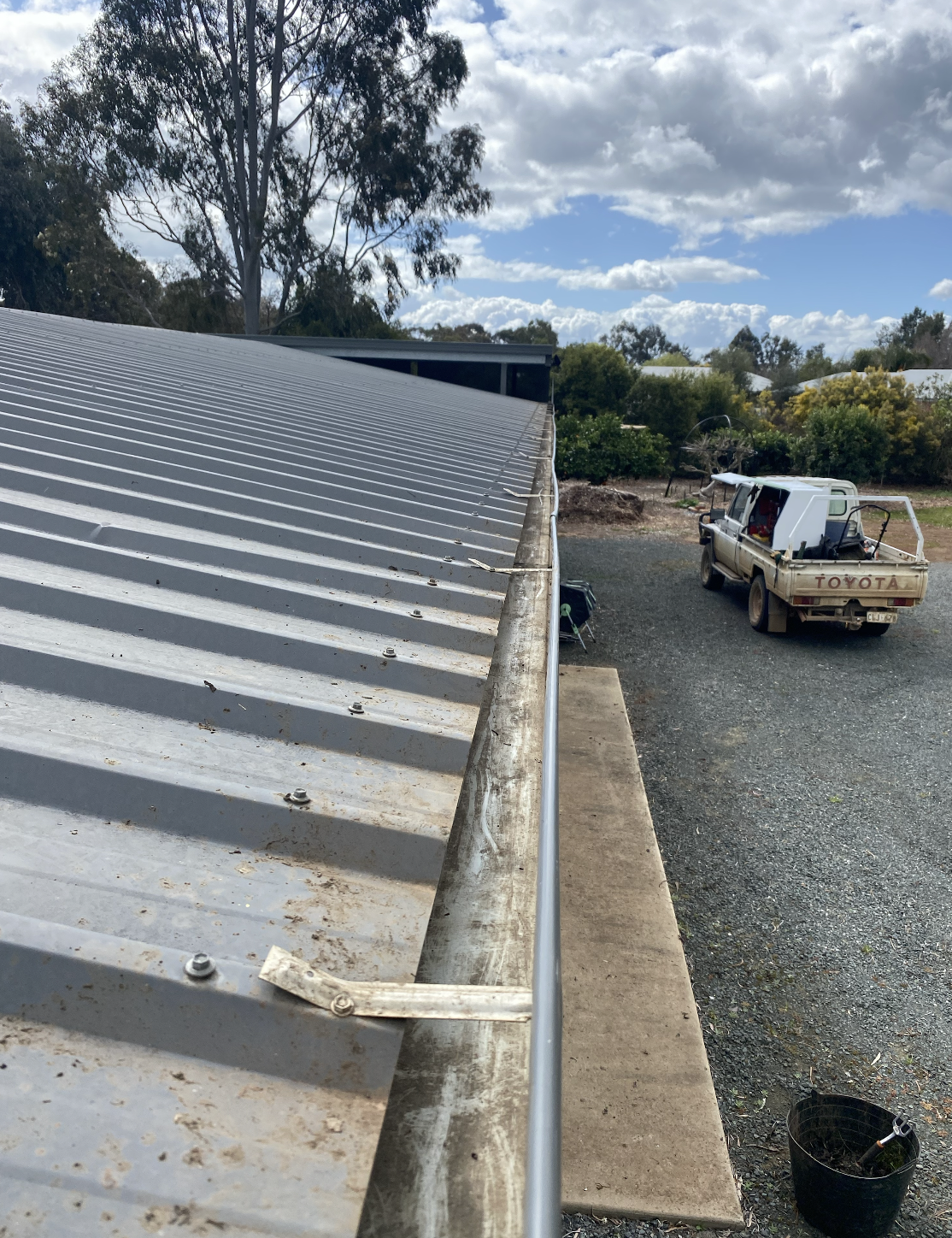 Close-up view of a metal roof with a gutter, with a pickup truck and a white vehicle in the background, trees, and a partly cloudy sky.