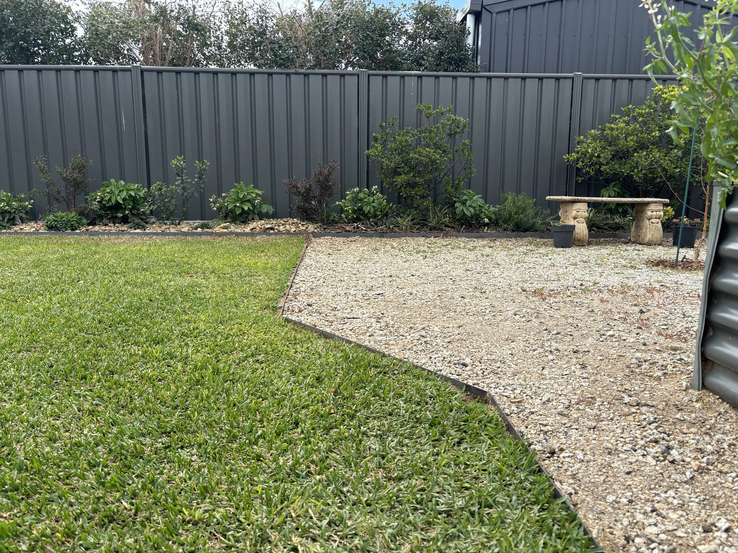 Garden backyard with a green lawn, a gravel path, a black metal fence, and small plants and bushes along the fence.
