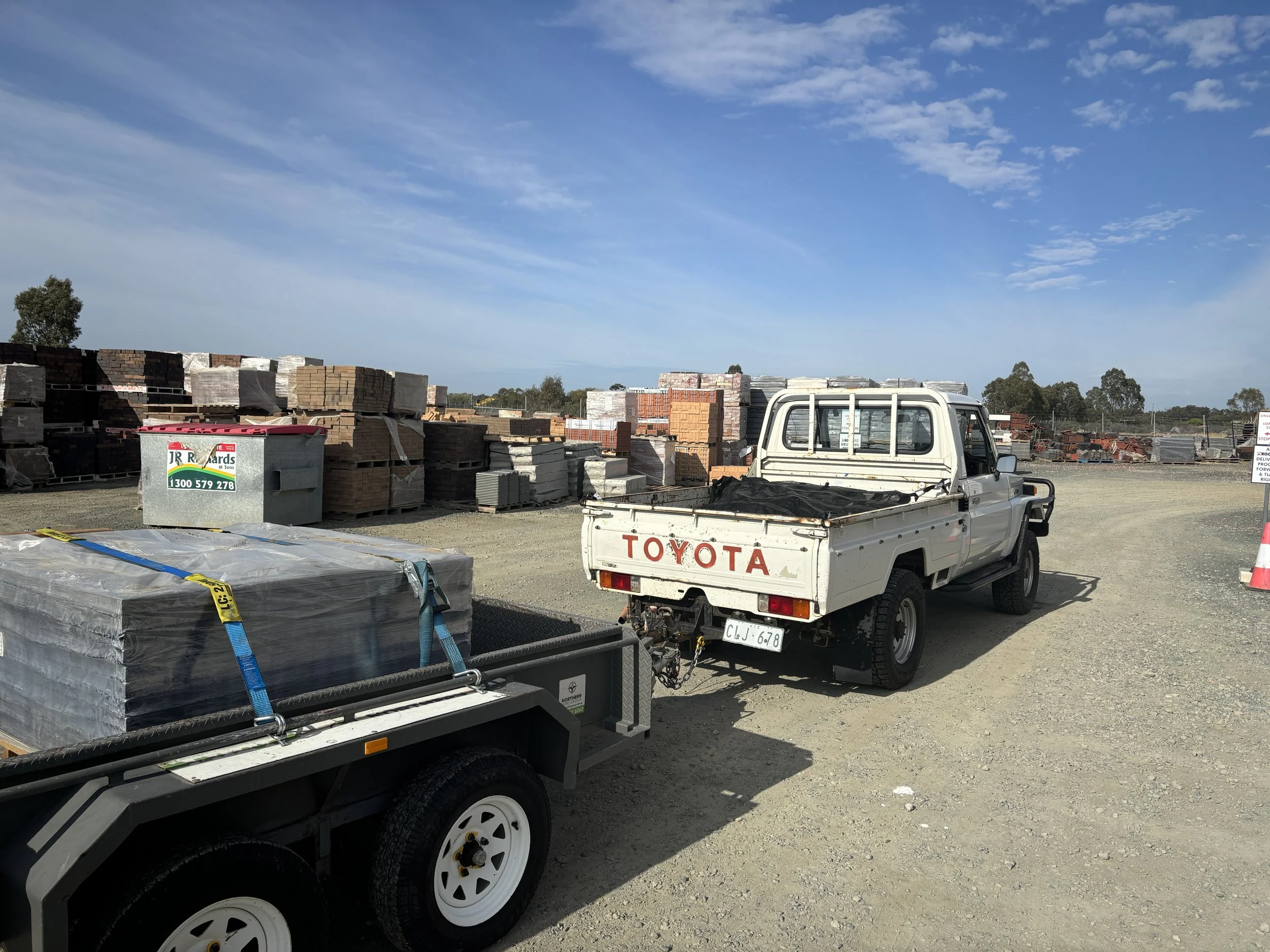 A white Toyota pickup truck parked in a construction materials yard with stacks of bricks and tiles in the background, under a partly cloudy sky.