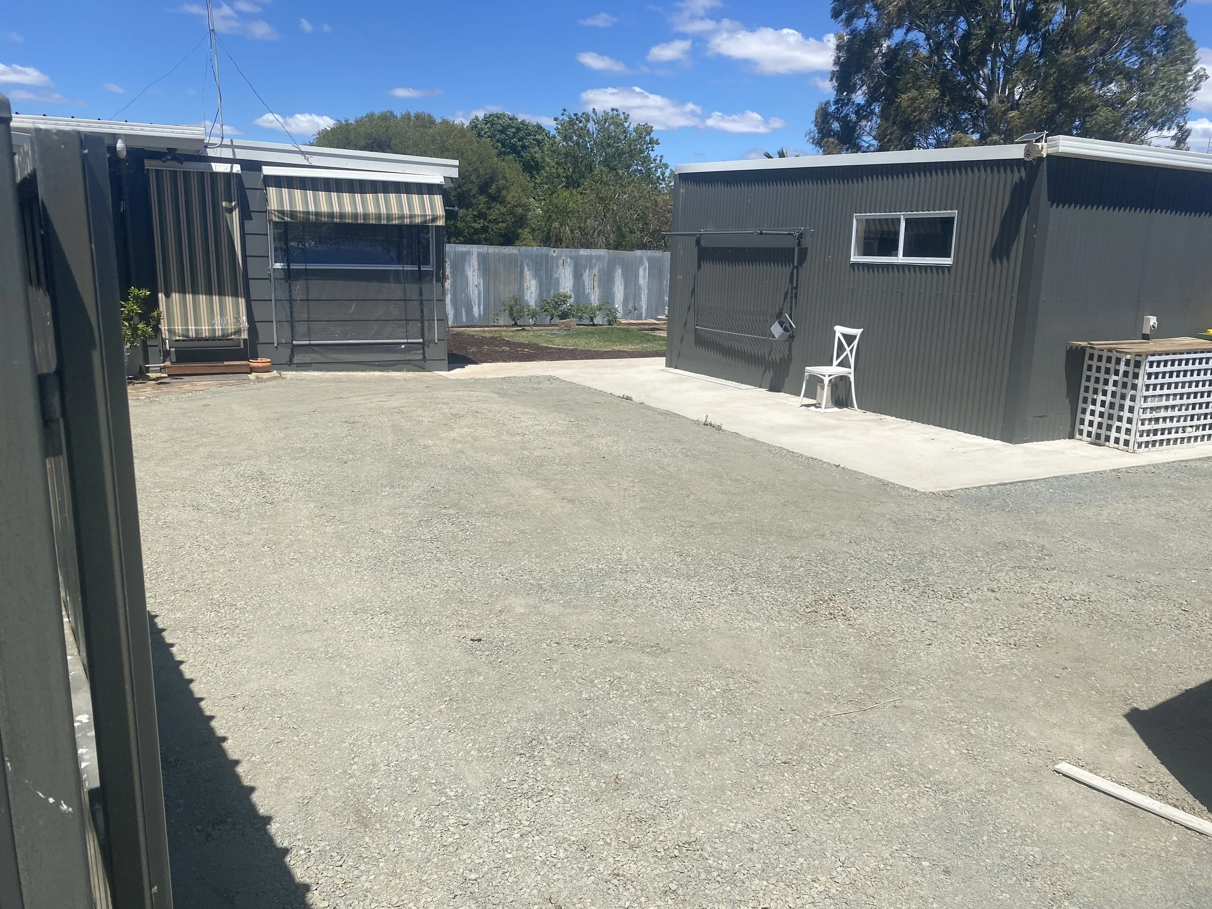 A small backyard with two gray metal sheds, one with a green and white striped awning, a white plastic chair, and a gravel ground under a blue sky with some clouds.