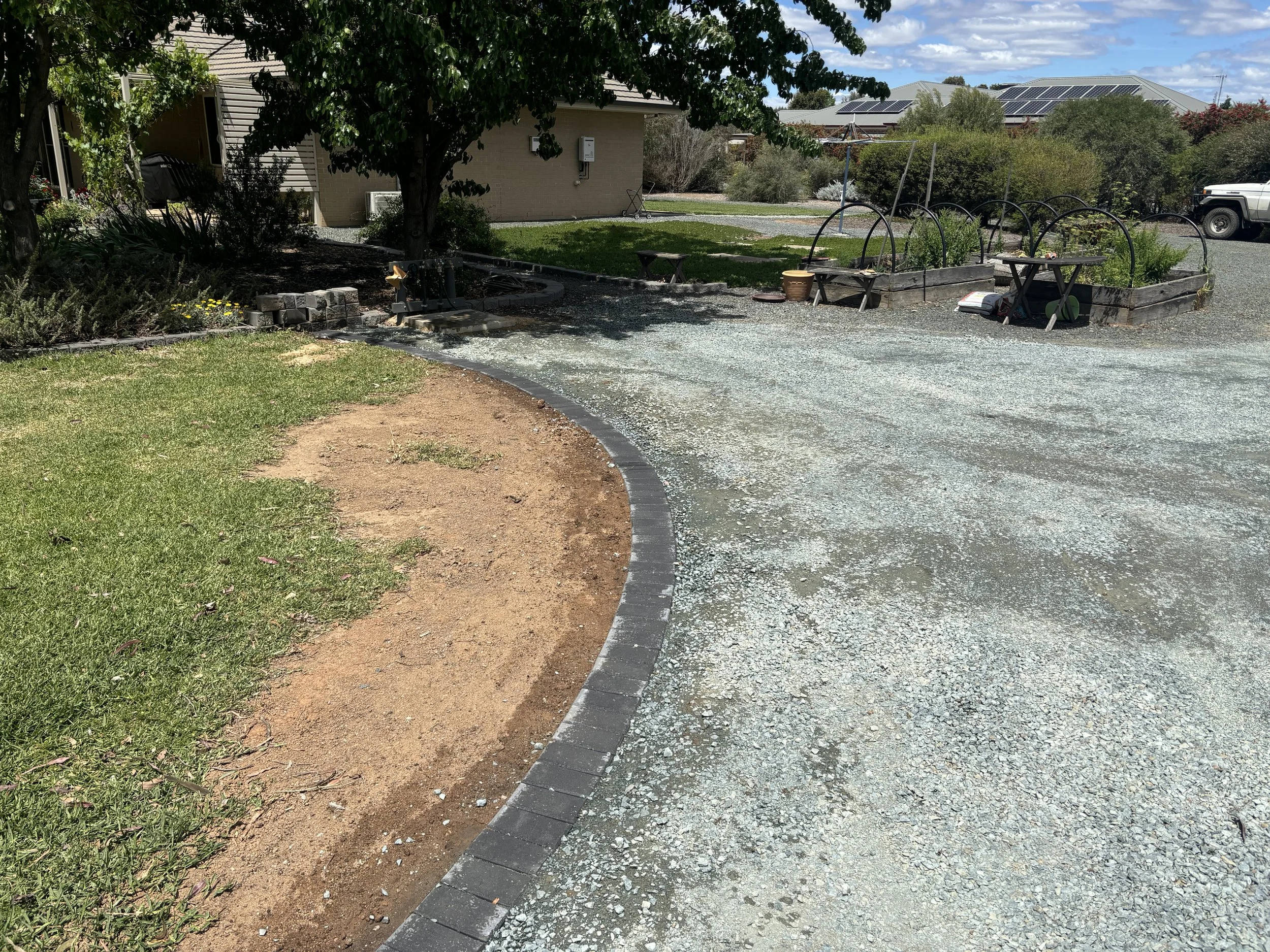 A yard with a gravel driveway, a green grass lawn, and patches of bare soil near the edge of the driveway. There are organized garden beds with plants and black arched supports. A house with tan siding and solar panels on the roof is in the background, along with a white truck parked on the gravel. A large tree provides shade over part of the yard.