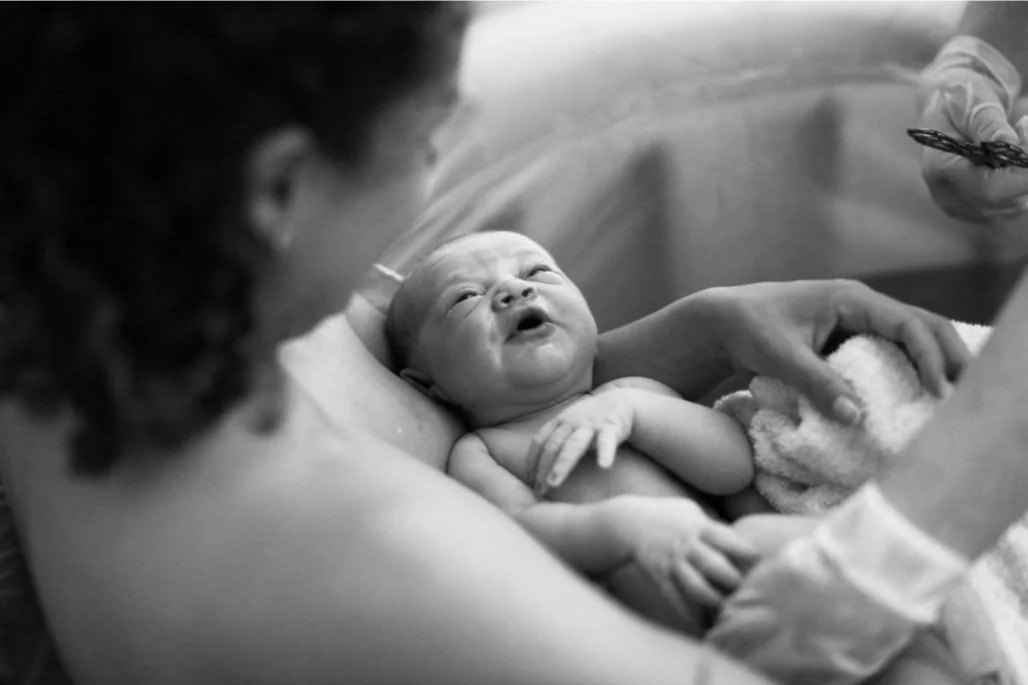 A newborn baby being held by a person, likely in a medical setting, while a healthcare worker prepares for a medical procedure or bath.