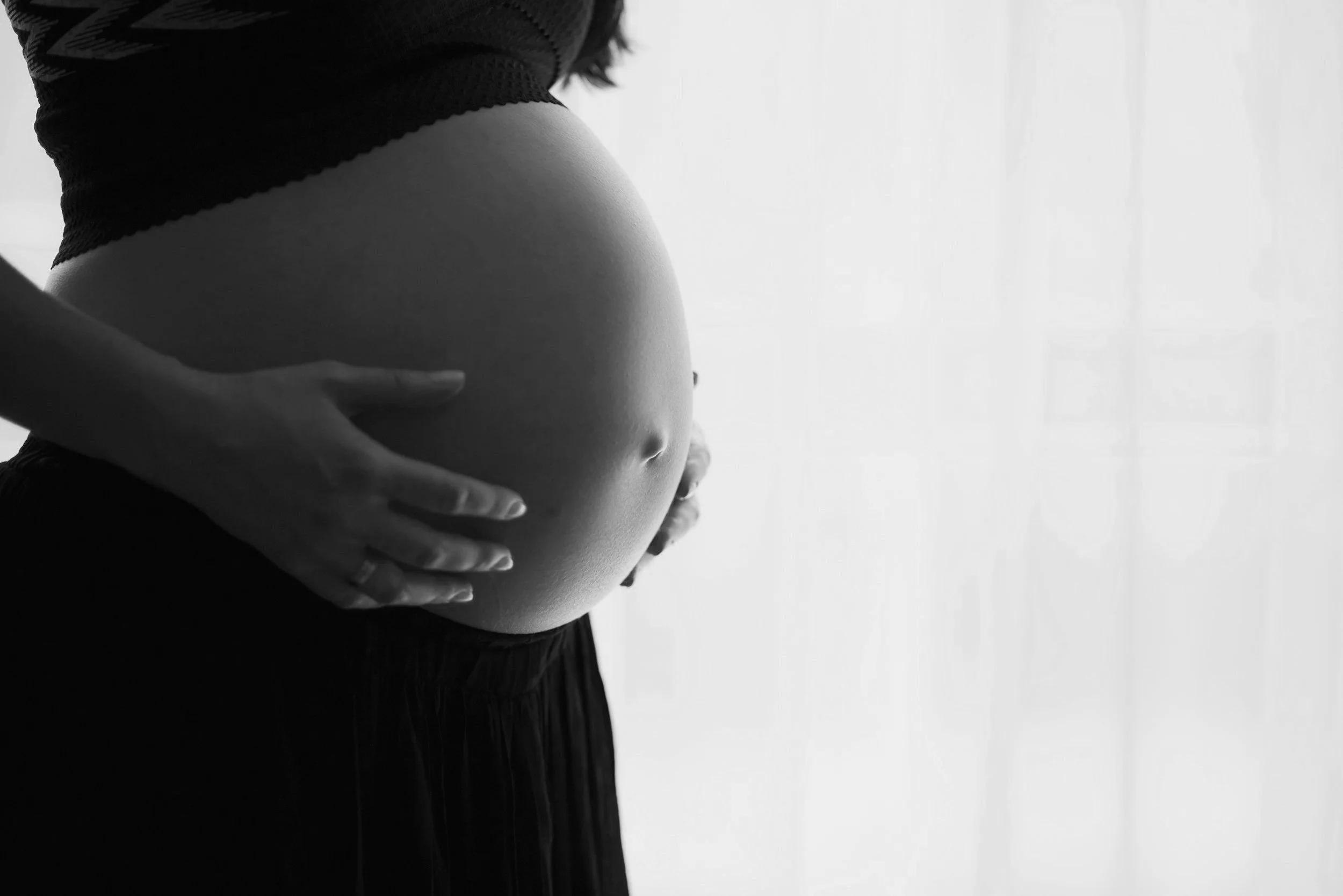 A pregnant person in a black and white photo gently resting their hands on their bare, rounded belly in front of a softly lit background.