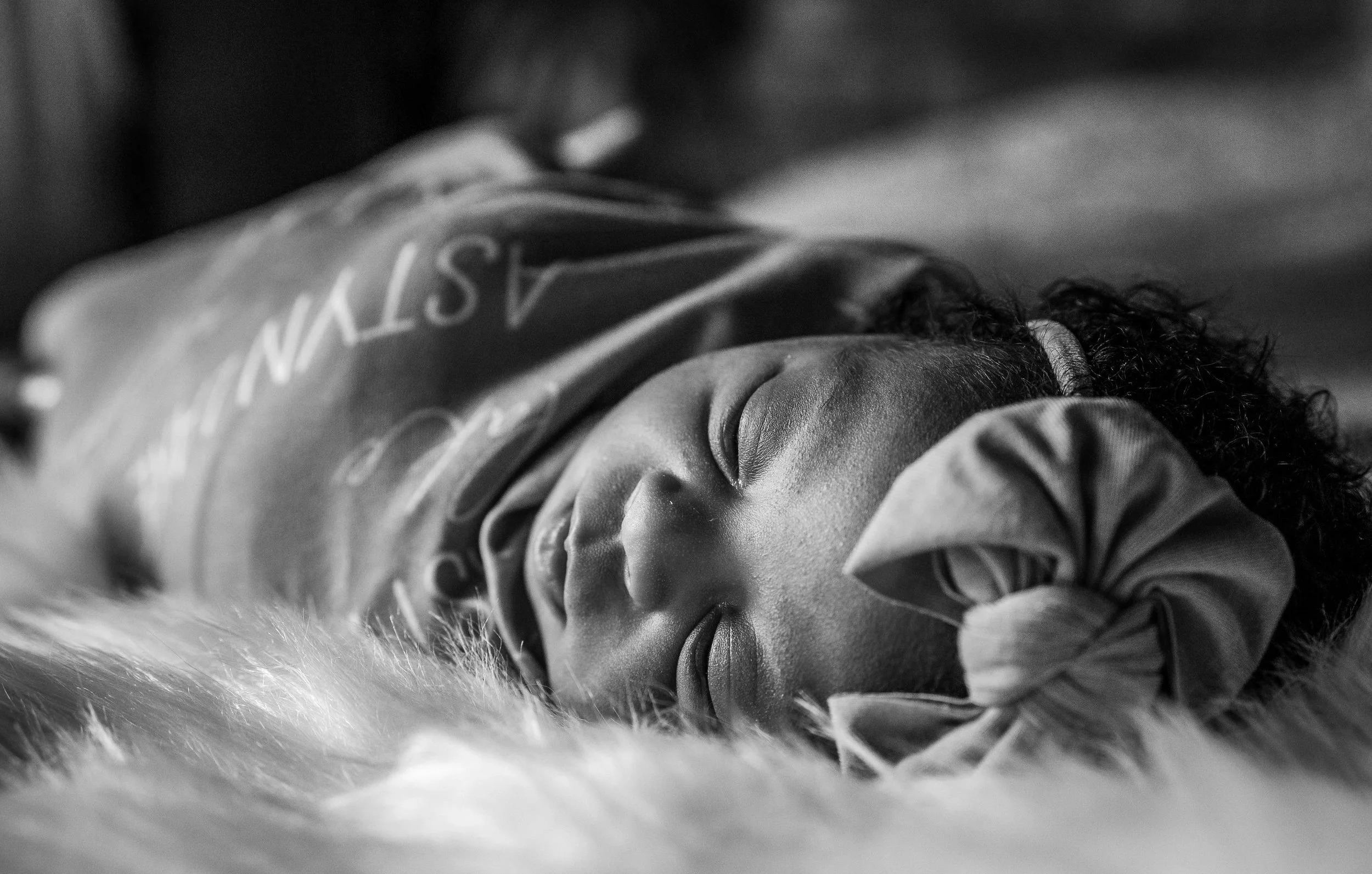 Black and white photo of a newborn baby sleeping on a fuzzy surface, wearing a headband with a bow.