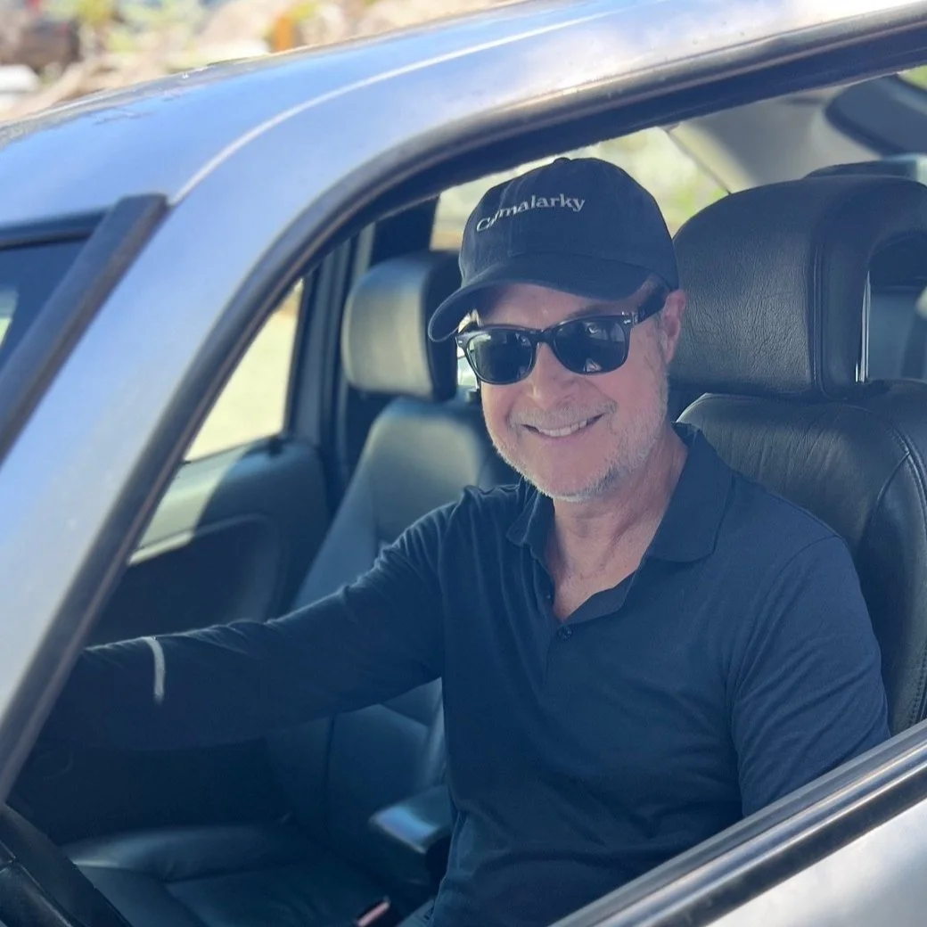 A smiling man wearing sunglasses and a black baseball cap sitting in the driver's seat of a blue car.