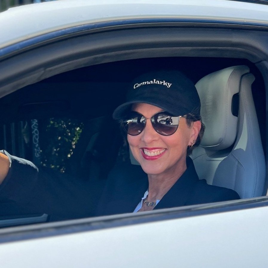 A woman with short hair, wearing sunglasses, a black cap with 'Carmalarky' embroidered on it, and a black outfit, is sitting in the driver's seat of a car, smiling.