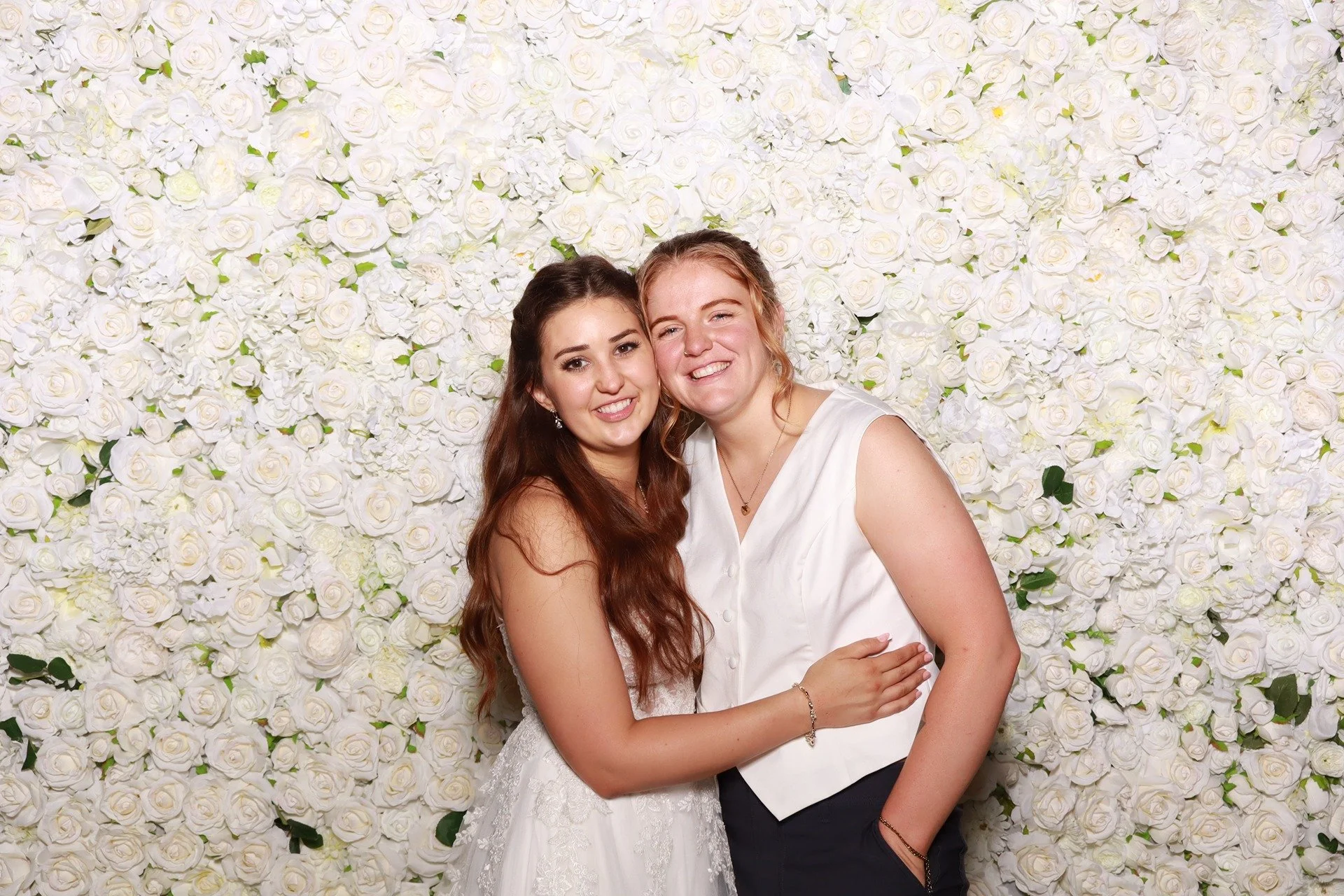  Bride smiling and hugging her partner in front of the white rose flower wall at Horning’s Hideout wedding photo booth. 