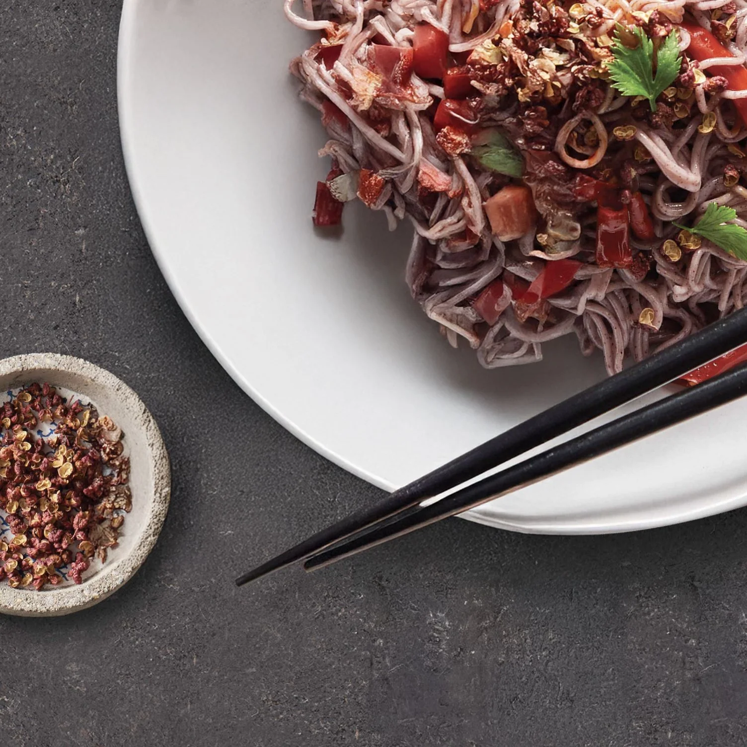 Plate of Koka Purple Noodles with red peppers and coriander garnish, served with chopsticks and pepper dish on dark surface – imported instant noodles supplied by Jetha Australian food importers