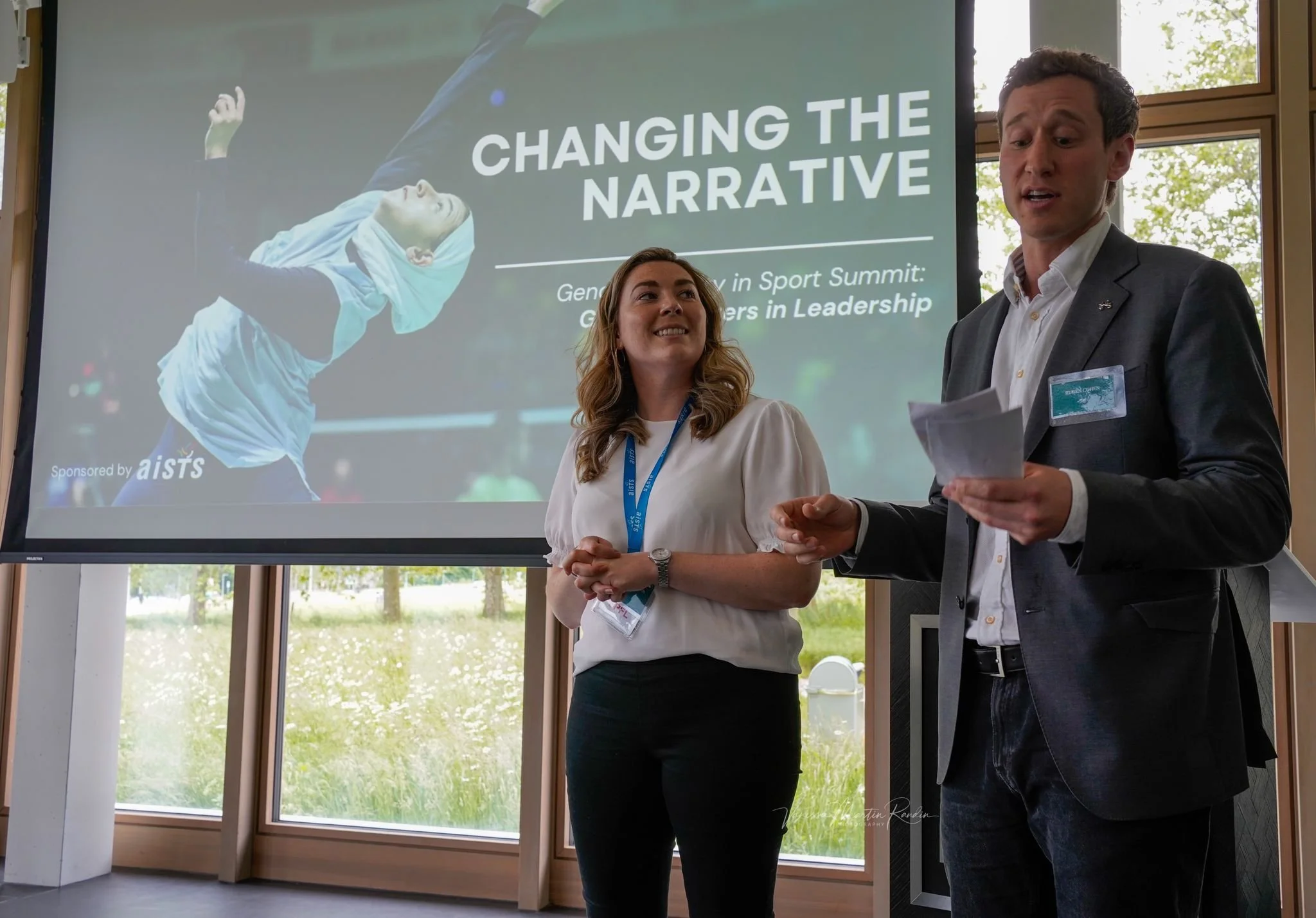 A man and a woman are standing in front of a presentation screen during a conference, with the screen displaying the title 'Changing the Narrative' and an image of a female badminton player in the background.