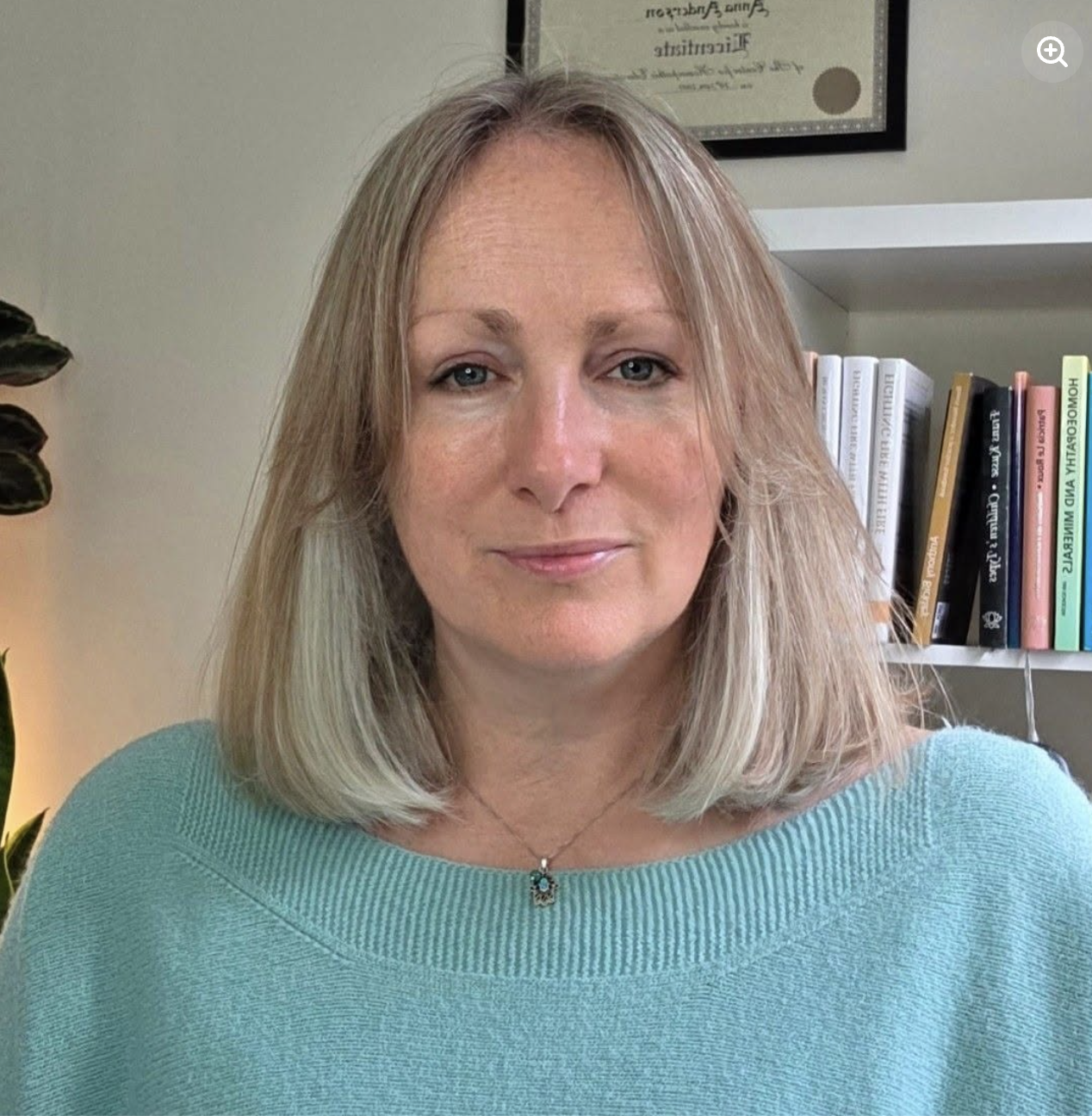 A woman with shoulder-length blonde hair and blue eyes wearing a light blue sweater and a necklace with a pendant, sitting in front of a bookshelf with white and black books and a framed certificate on the wall.