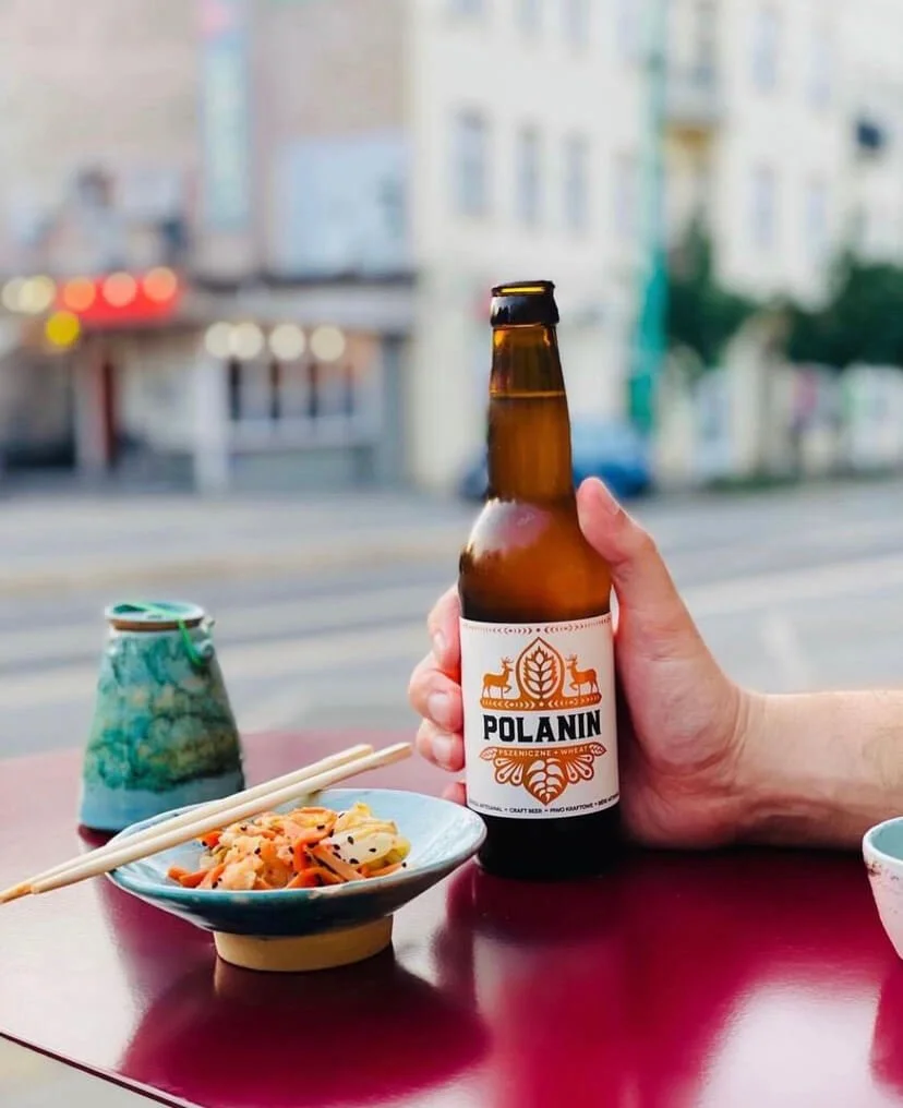 Person holding a bottle of Polanin beer on an outdoor table with a bowl of Asian food, a pair of chopsticks, and a ceramic cup, with city buildings in the background.