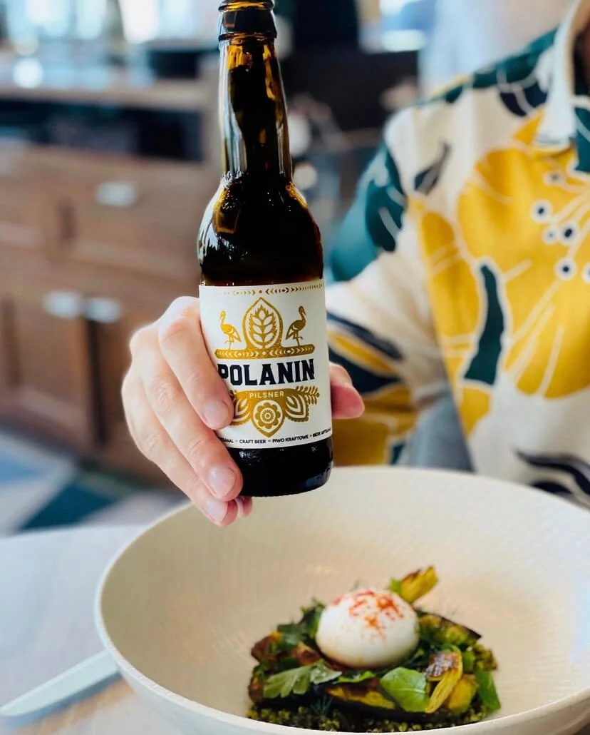 A person holding a bottle of Polanin Pilsner beer over a dish with greens, vegetables, and a poached egg in a white bowl.