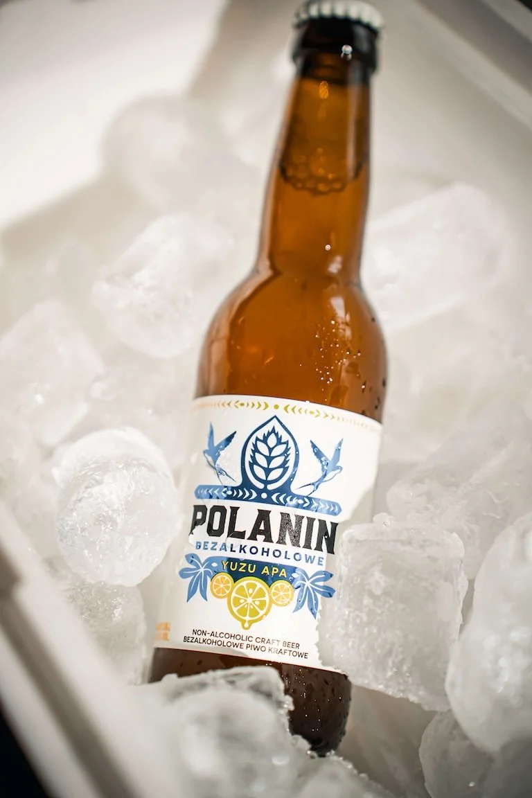 A brown bottle of Polanin non-alcoholic craft beer with a label featuring lemons, hops, and two birds, resting in an ice-filled container.