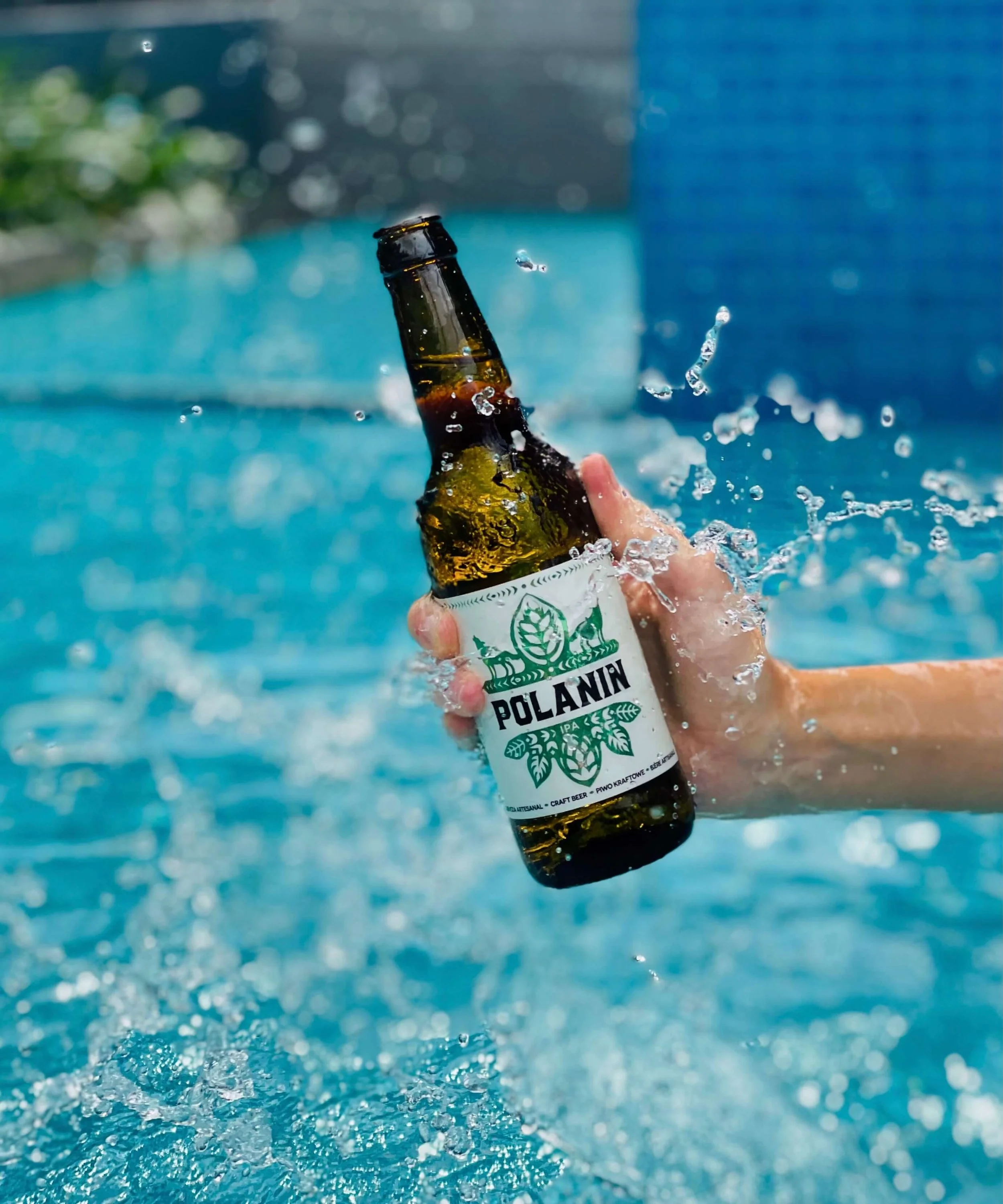 A person is opening a bottle of Polanin craft beer in a swimming pool, with water splashing around.