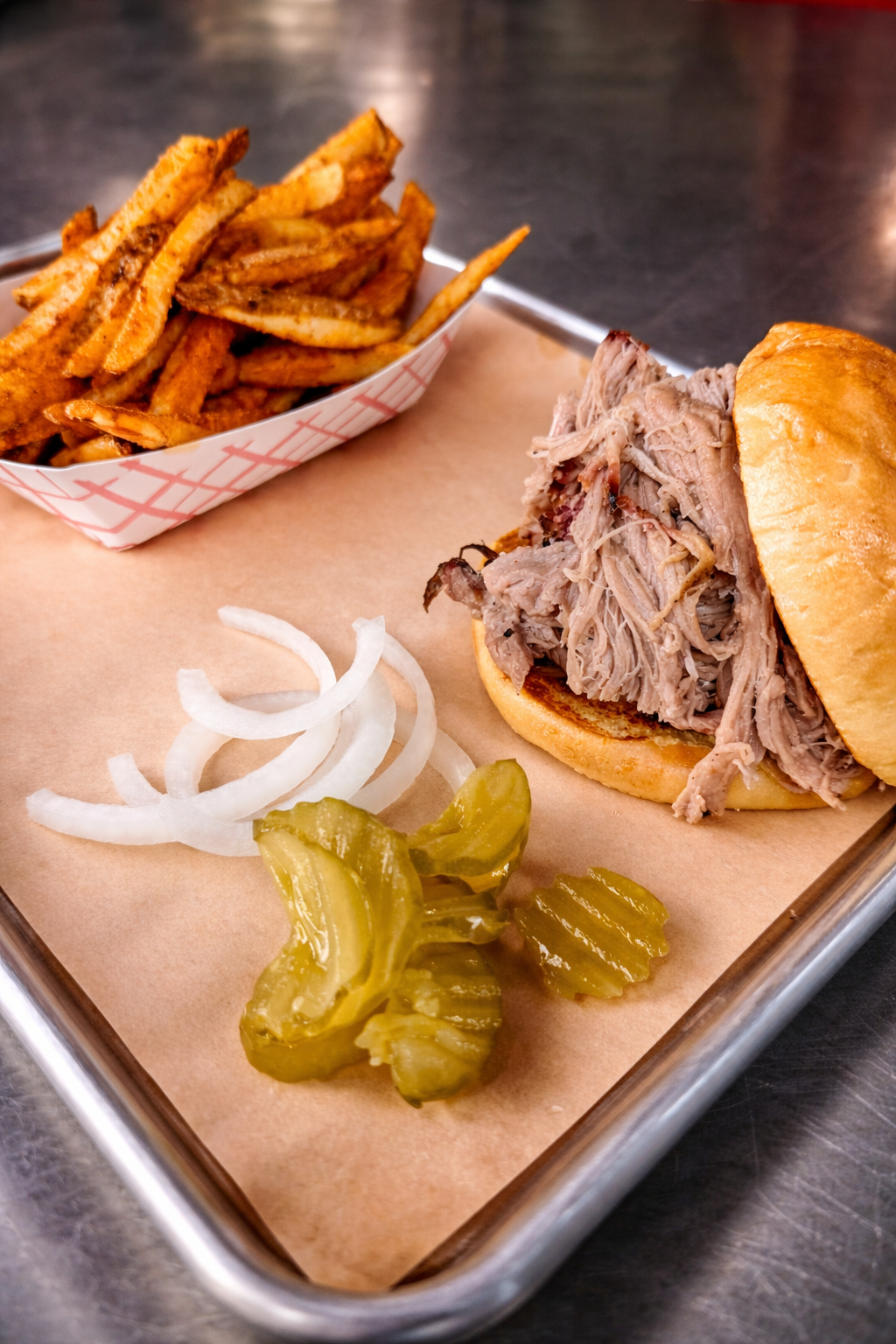 Moist flavorful Pulled pork sandwich with pickles, sliced onions, and a side of seasoned fresh cut French fries in a paper boat, served on a tray with butcher paper.