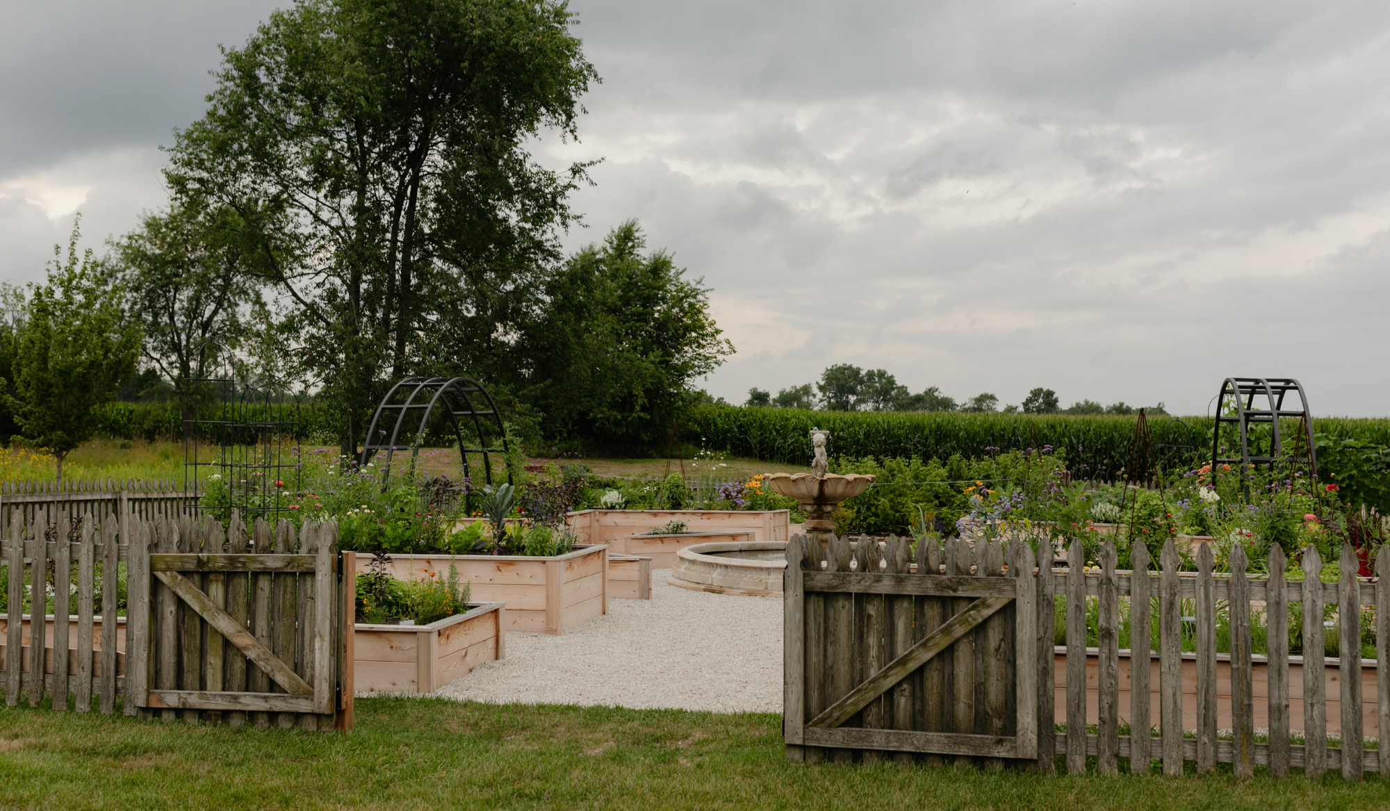 Open wooden gates leading into the LaRocque Farms kitchen garden, with cedar raised beds, flowers, arch trellises, and the central fountain beyond.