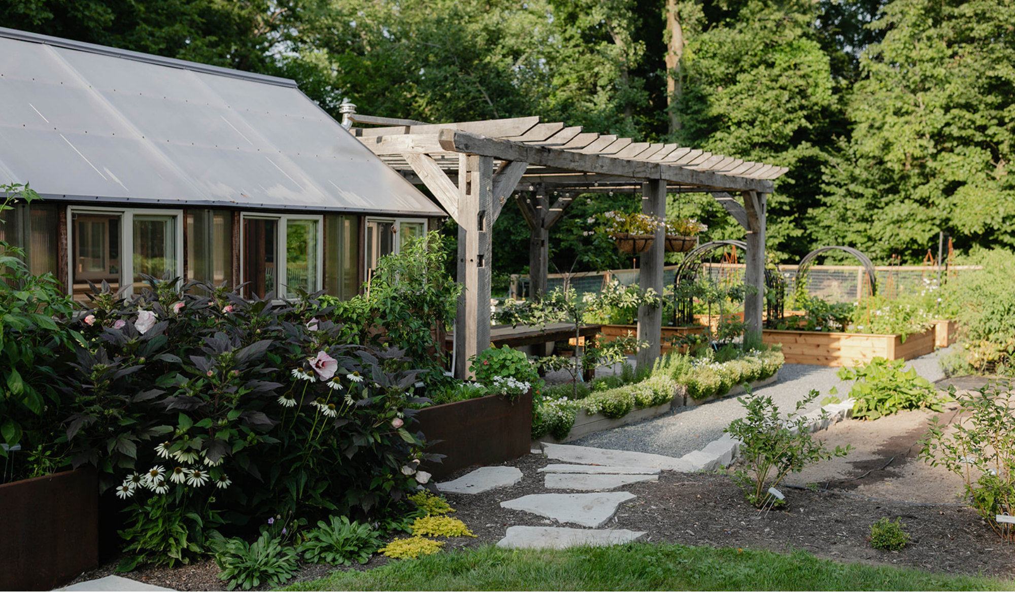 Side view of the Yoderbilt greenhouse and reclaimed timber pergola, connected by bluestone stepping stones and gravel pathways surrounded by lush plantings.