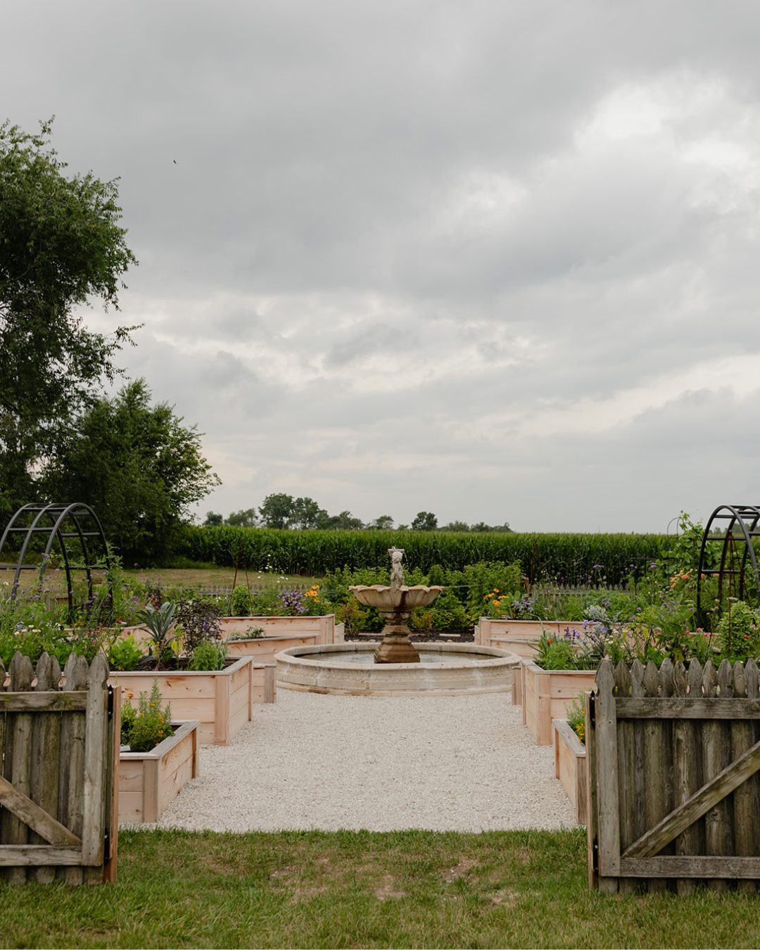 Open wooden gates framing the central fountain, cedar raised beds, colorful plantings, and black arch trellises of the LaRocque Farms kitchen garden.