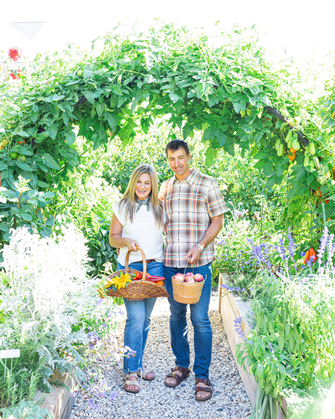 Katie Oglesby and her husband Matt standing beneath a tomato-covered arch trellis with baskets of fresh garden harvests at The Edible Acre.