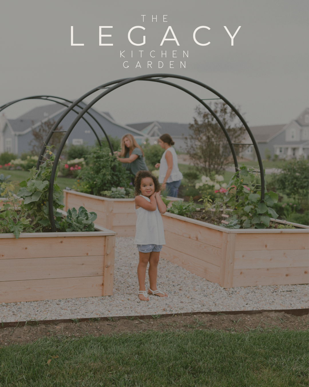 Young girl standing beneath the double arch trellises in The Legacy Garden, surrounded by cedar raised beds while family members tend the garden behind her.