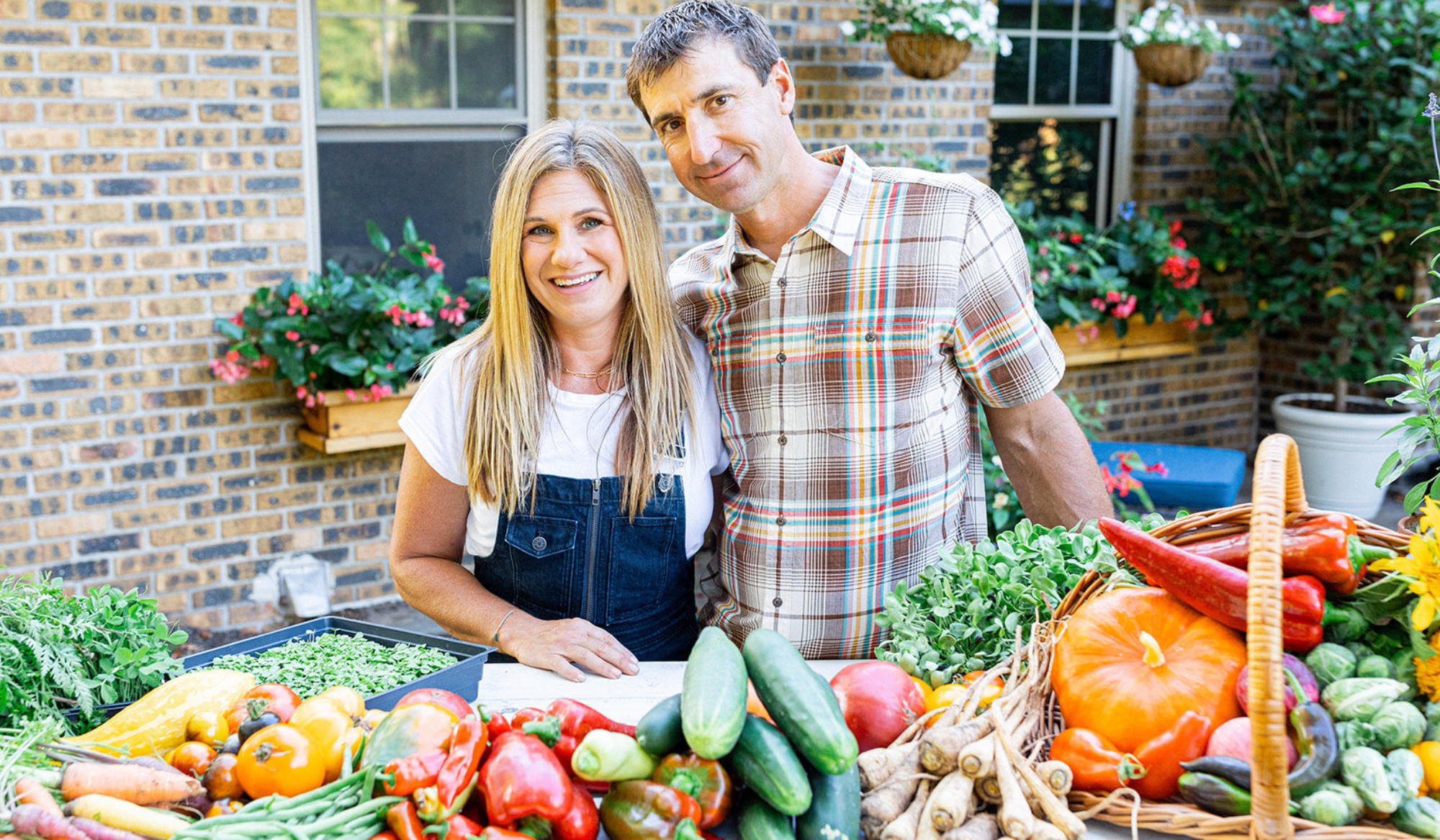Katie and Matt Oglesby standing behind a colorful harvest of homegrown vegetables, including tomatoes, peppers, cucumbers, squash, carrots, and pumpkins.