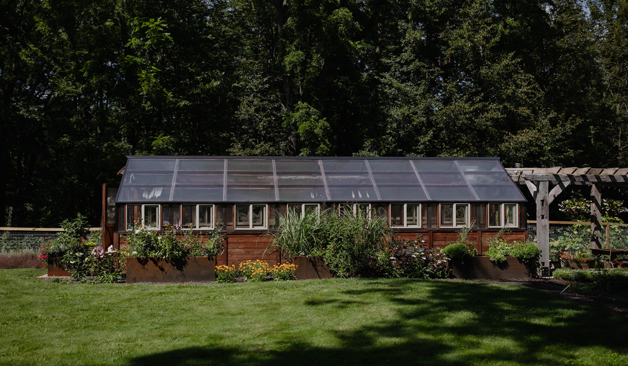 Yoderbilt greenhouse at The Edible Acre in southern Wisconsin, surrounded by corten steel planters, flowering plants, and landscaped garden beds.