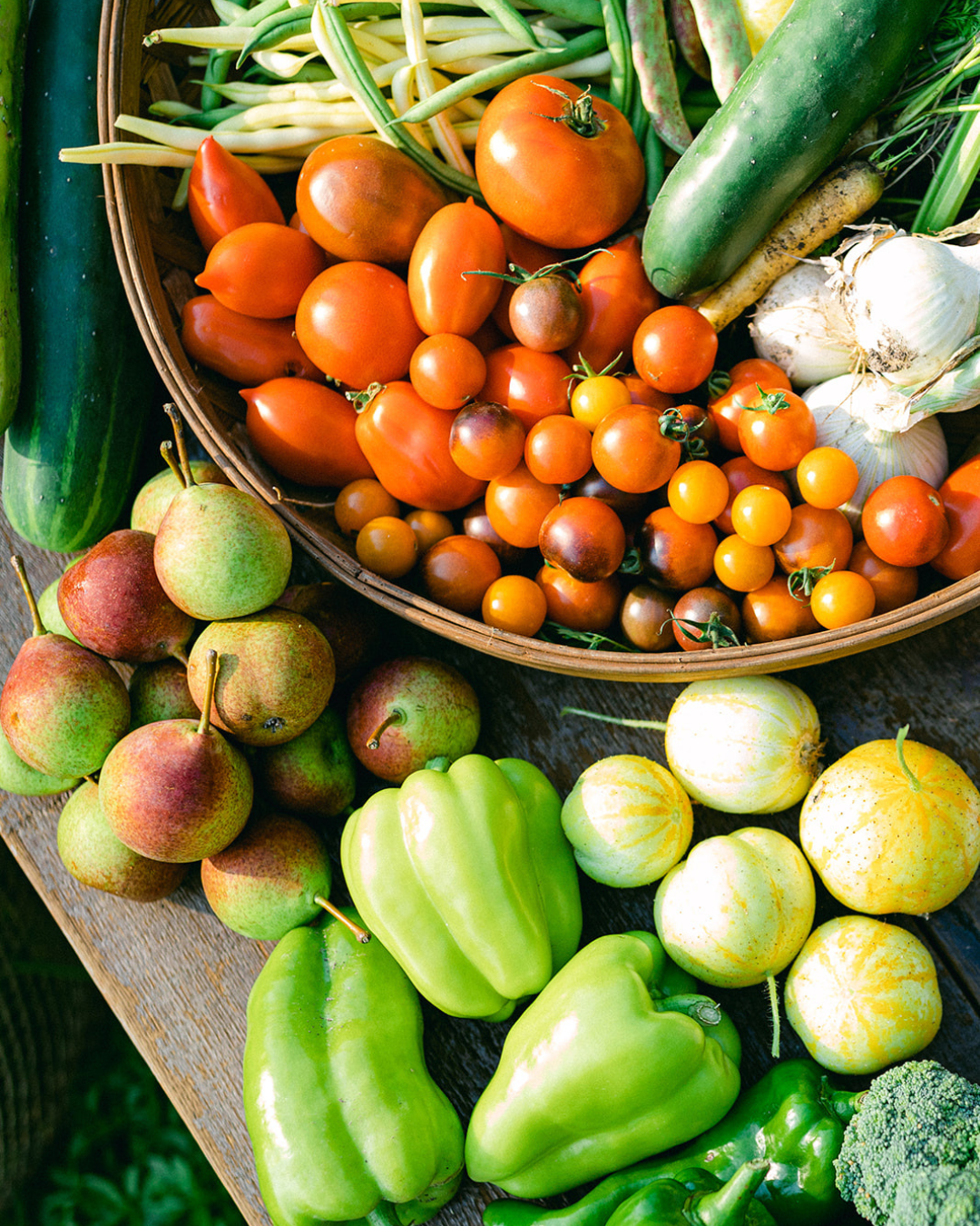 Freshly harvested tomatoes, pears, cucumbers, peppers, beans, garlic, broccoli, and melons arranged on a wooden table.