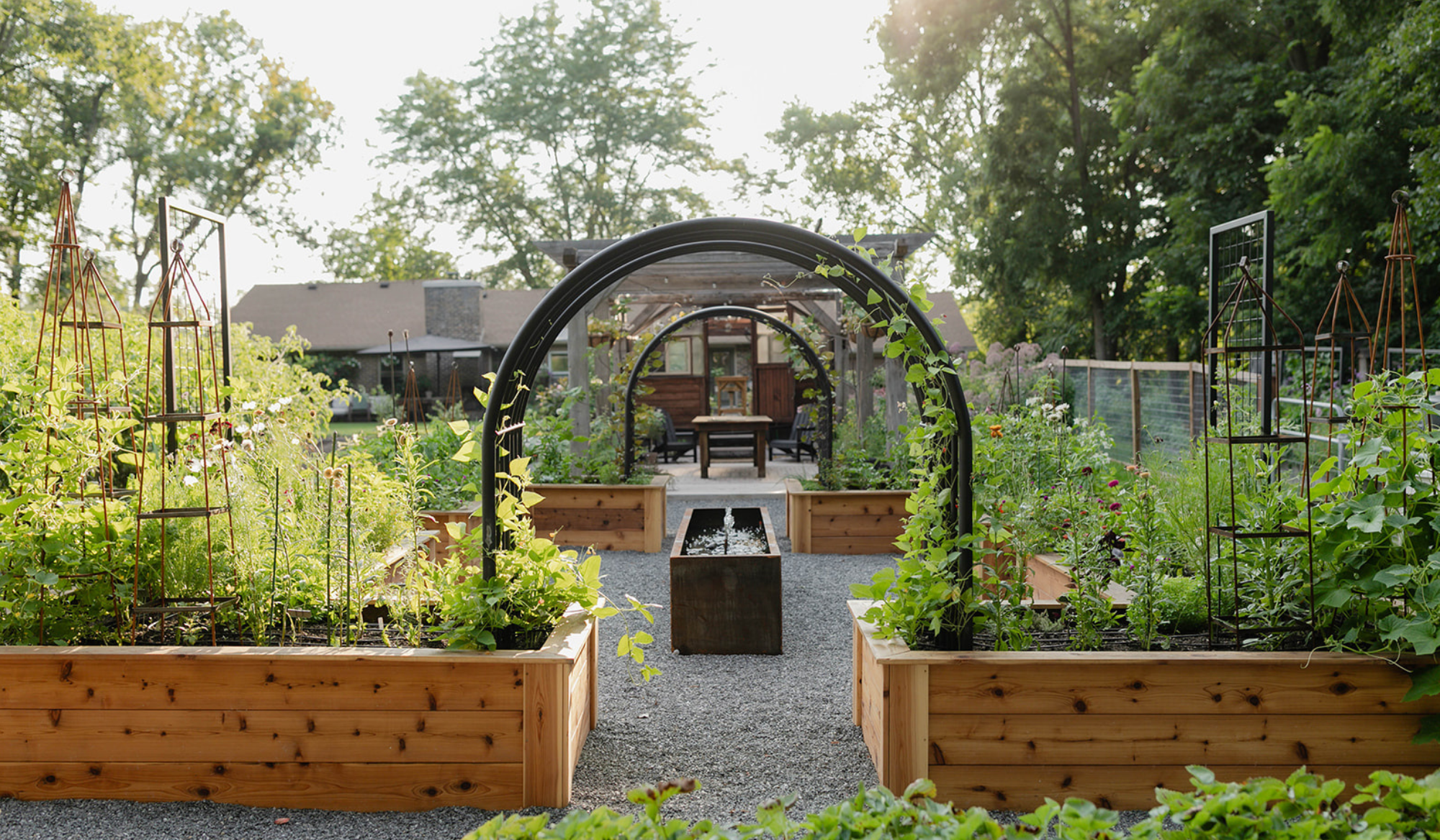 The new kitchen garden at The Edible Acre, featuring cedar raised beds, black arch trellises, a custom corten steel water feature, and a pergola beyond.