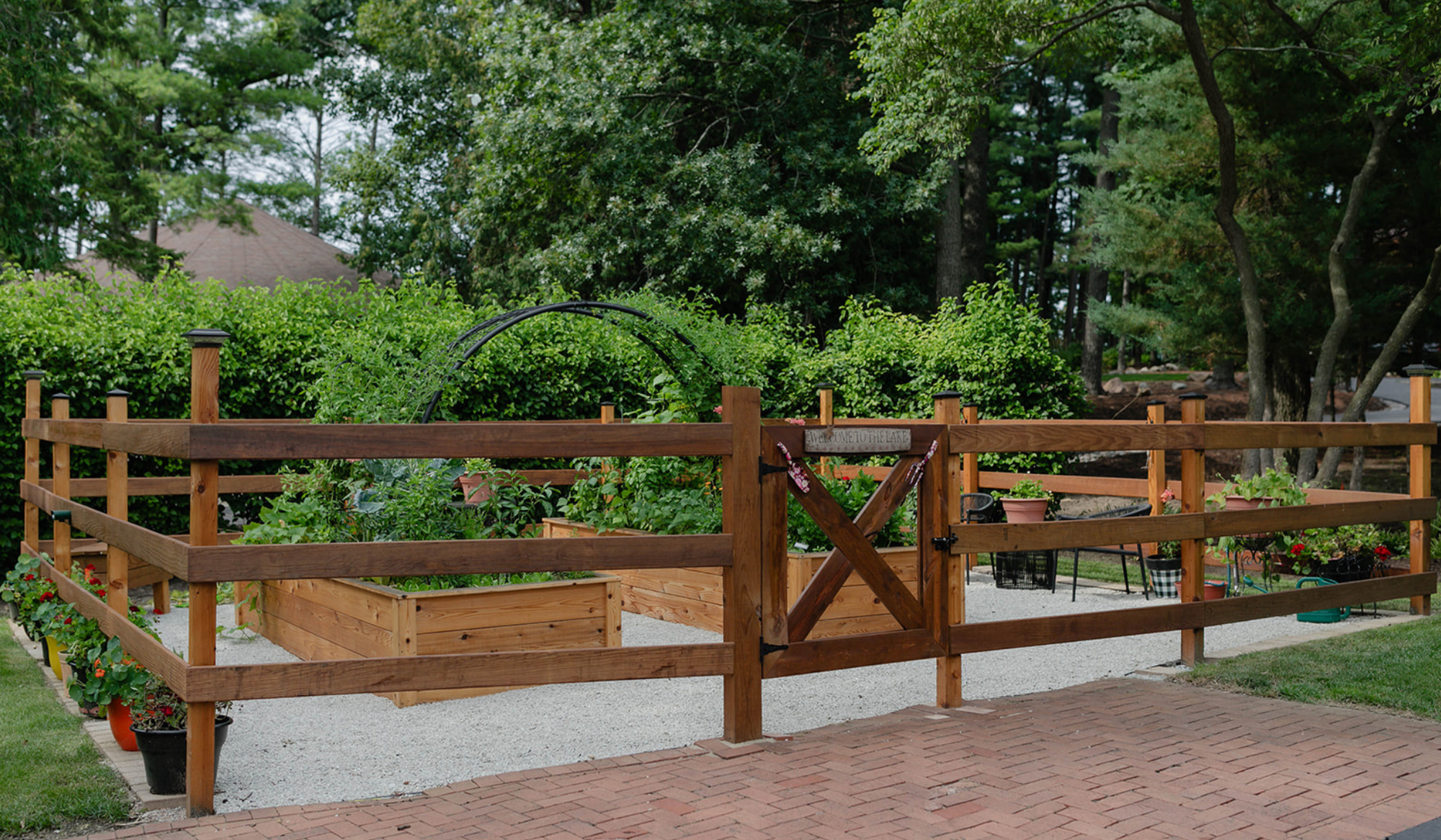 The Hawks Garden enclosed by a wood fence and gate, with cedar raised beds, white gravel pathways, a custom arch trellis, and sitting area.
