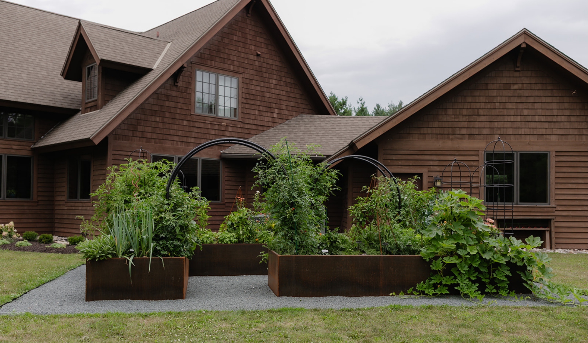 Corten steel kitchen garden with vegetables, flowers, black trellises, and gray stone pathways beside a cedar-sided home