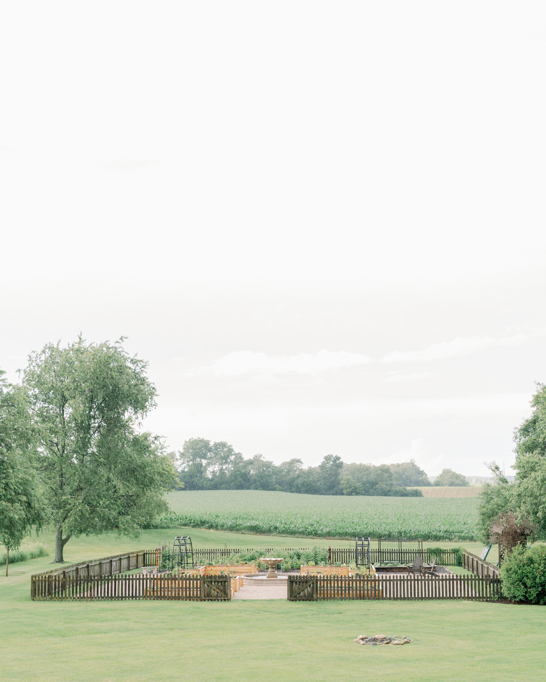 LaRocque Farms kitchen garden surrounded by lawn and farmland, with a fenced potager, cedar raised beds, black trellises, and central fountain.