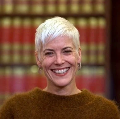 A woman with short white hair and earrings, smiling in front of a background of books.