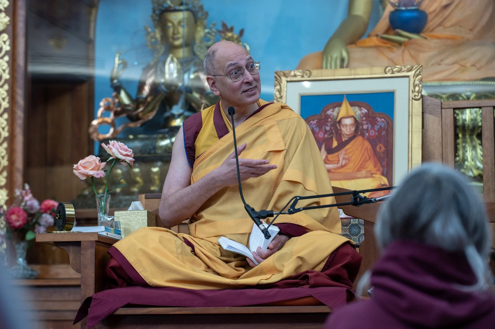A man wearing traditional Buddhist robes sits cross-legged on a cushion, speaking into a microphone, with an open book in his lap. There are floral arrangements and a box of tissues on a table nearby. In the background, there are colorful religious statues and a framed photograph or painting of an elderly Buddhist teacher.