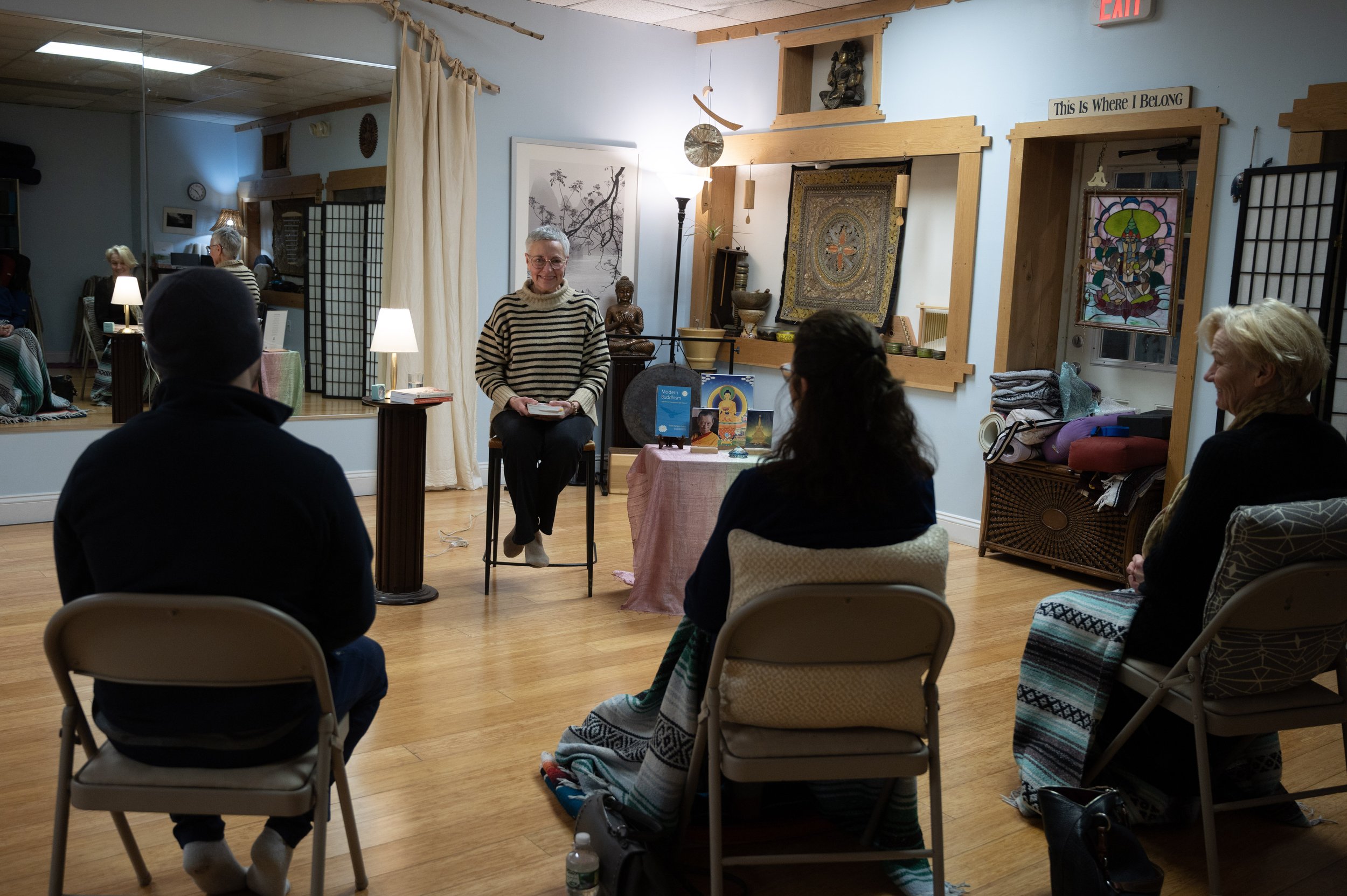 A woman with gray hair, glasses, and a striped sweater sits on a stool, smiling, in front of a group of people attending a presentation or discussion in a cozy room decorated with spiritual and Asian art.