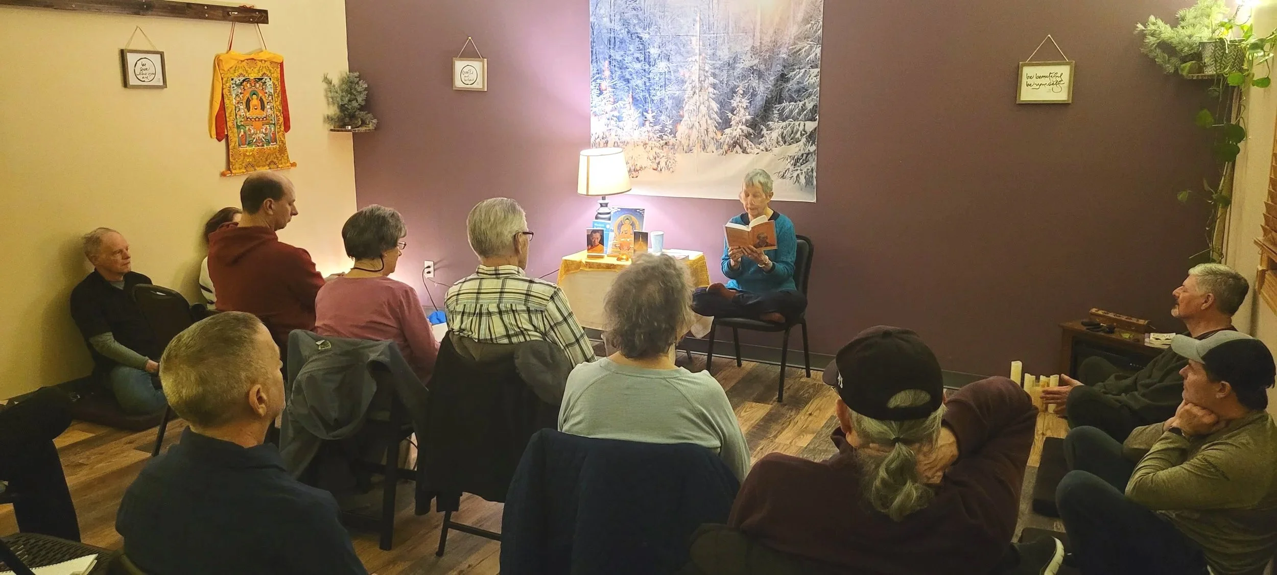 A woman is sitting on a chair, reading from a book to a group of people in a cozy room with purple and yellow walls, a winter-themed wall hanging, a lamp, potted plants, and framed pictures.