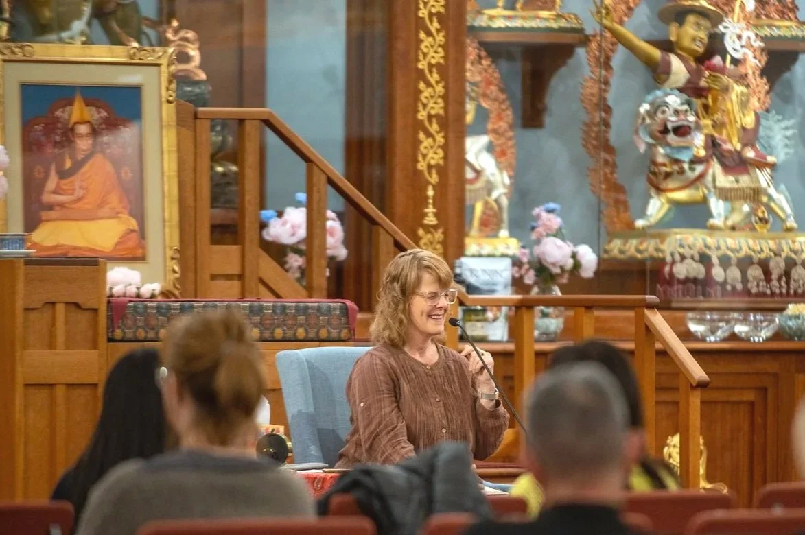 Woman with glasses smiling and speaking into a microphone in a room with ornate Asian artwork and decorations, including a portrait of a religious figure and colorful statues of dragons and a figure playing a musical instrument.