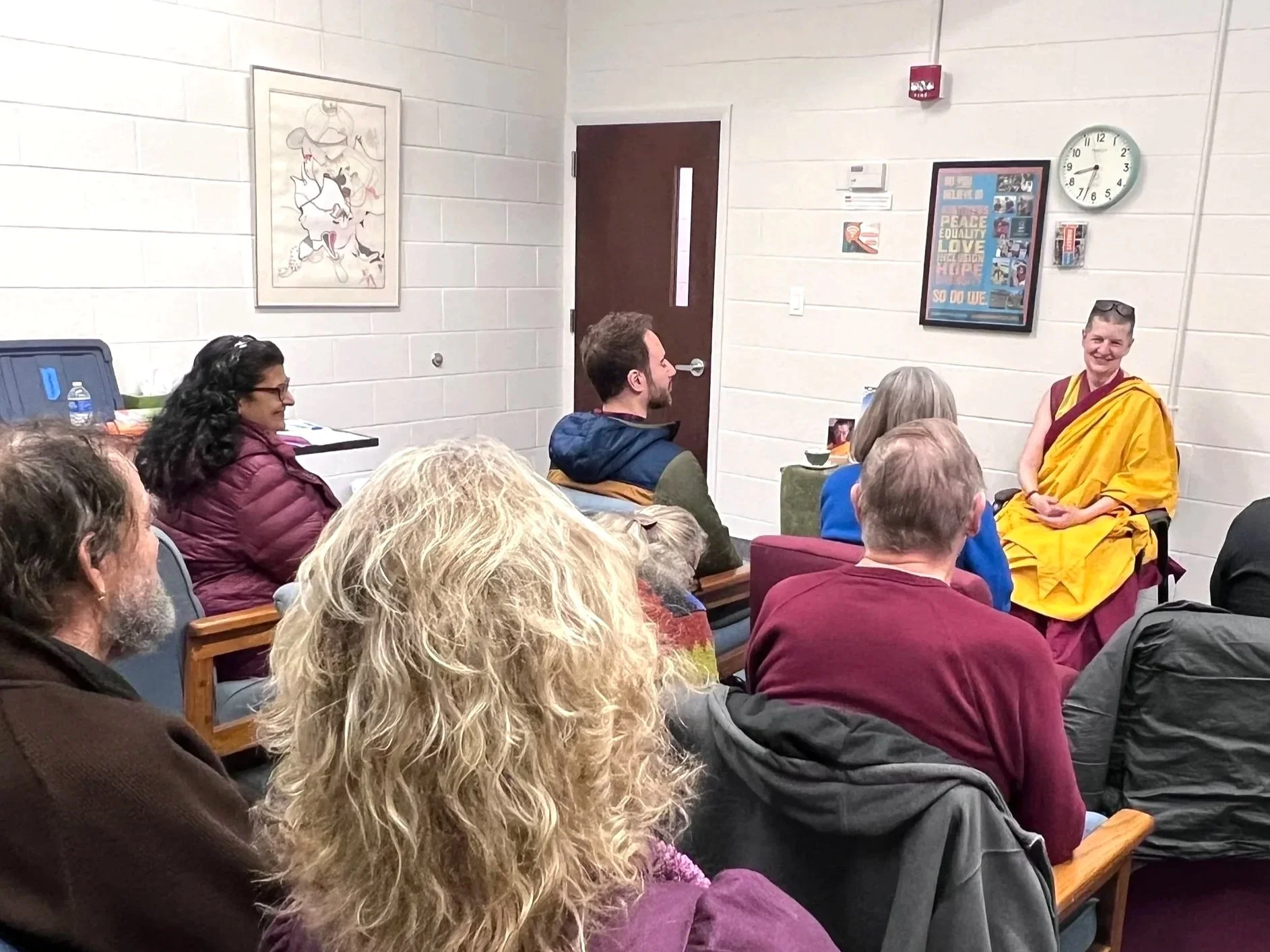 A group of people gathered in a room, listening to a person dressed as a Buddhist monk seated at the front. The room has light-colored walls, and is located in Binghamton, New York.
