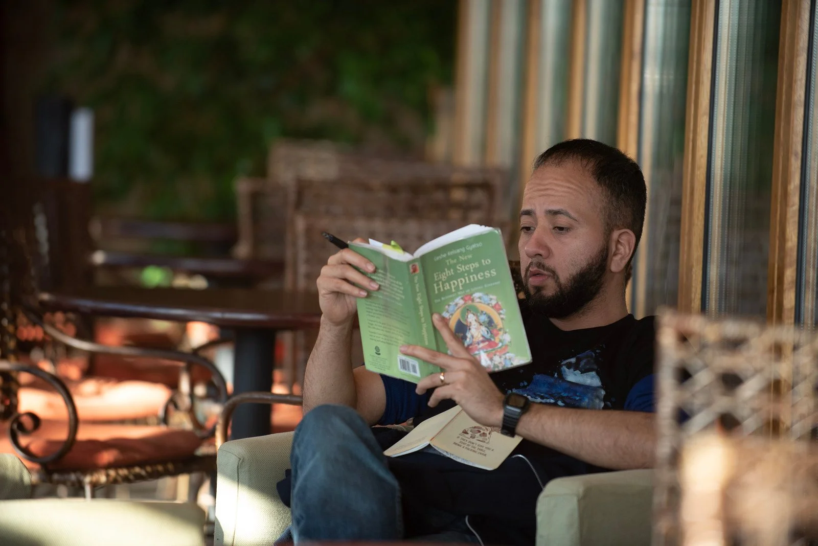 A man with a beard reading a book titled 'Eight Steps to Happiness' while sitting on a beige armchair near a window with wooden blinds.