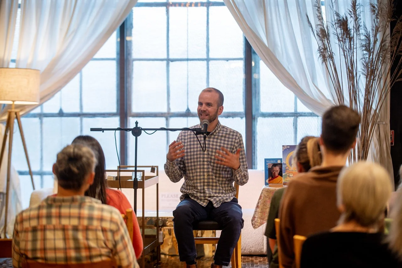 Kadam Kyle into a microphone while sitting on a stool in front of an audience in a cozy room with large windows and draped curtains.