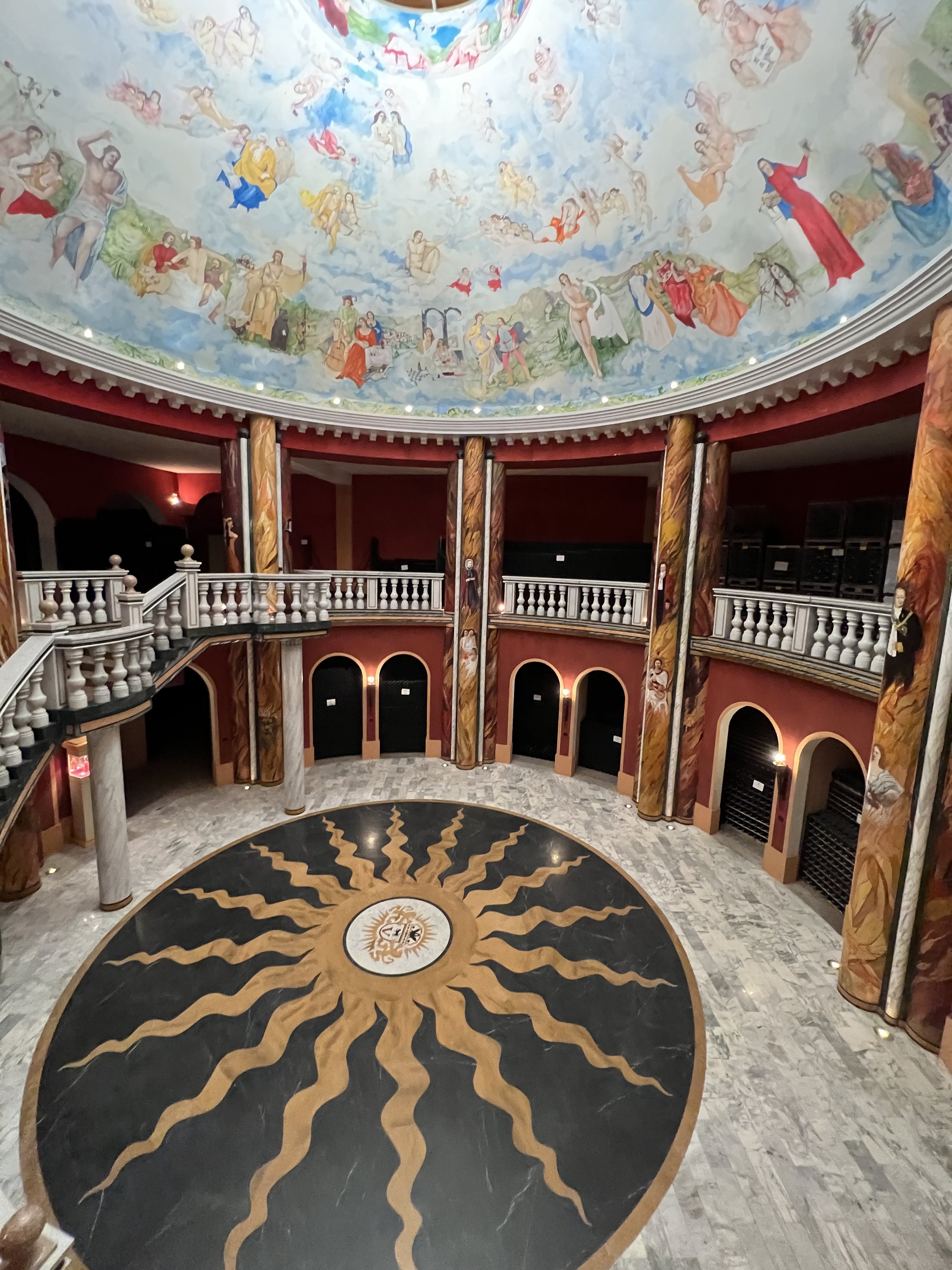 Interior view of a circular hall with a painted domed ceiling depicting numerous mythological and historical figures in clouds, a decorative marble floor with a sun motif, and ornate columns with painted designs supporting the upper balcony.