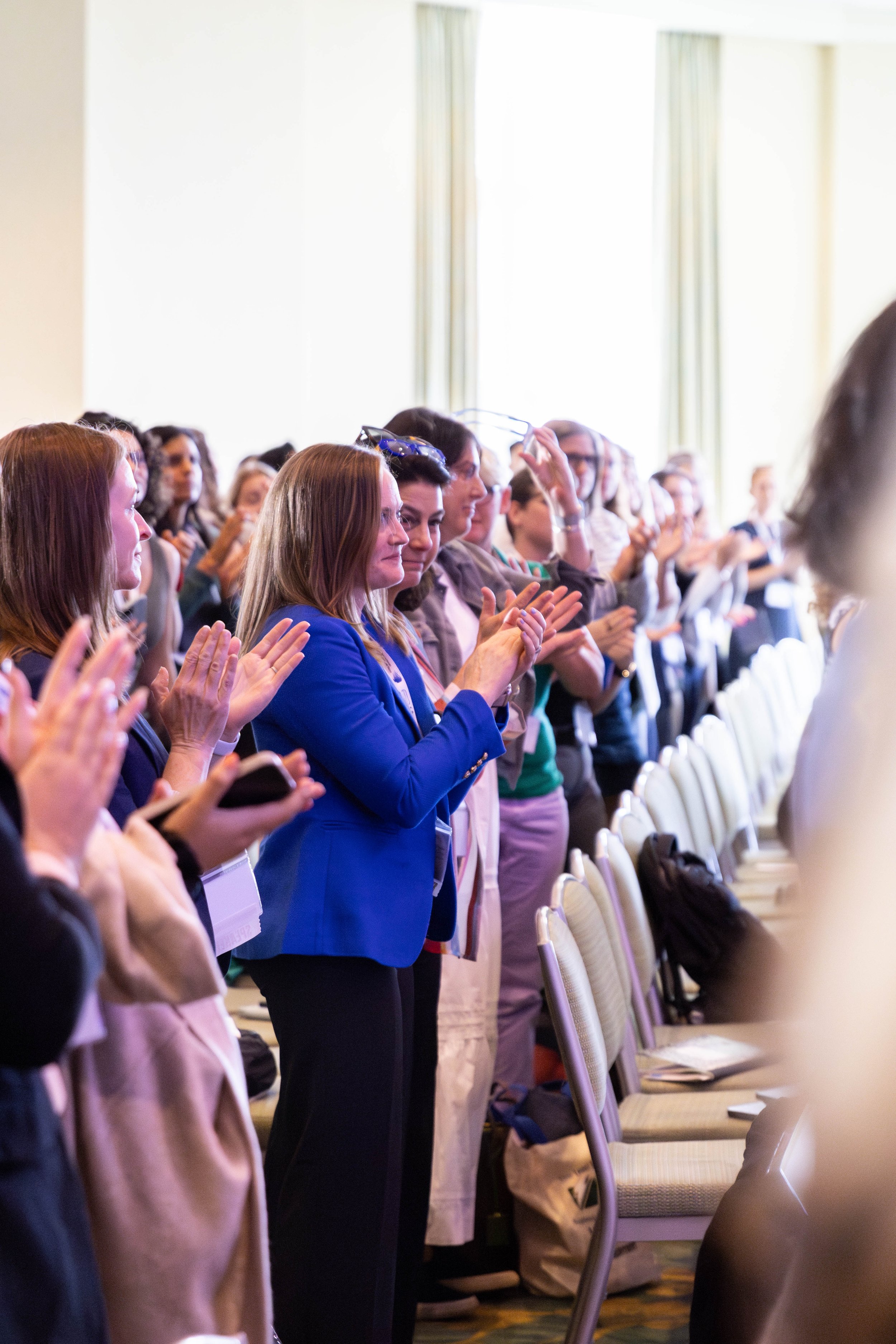 A group of diverse women standing and clapping in a conference room with bright light coming from large windows.